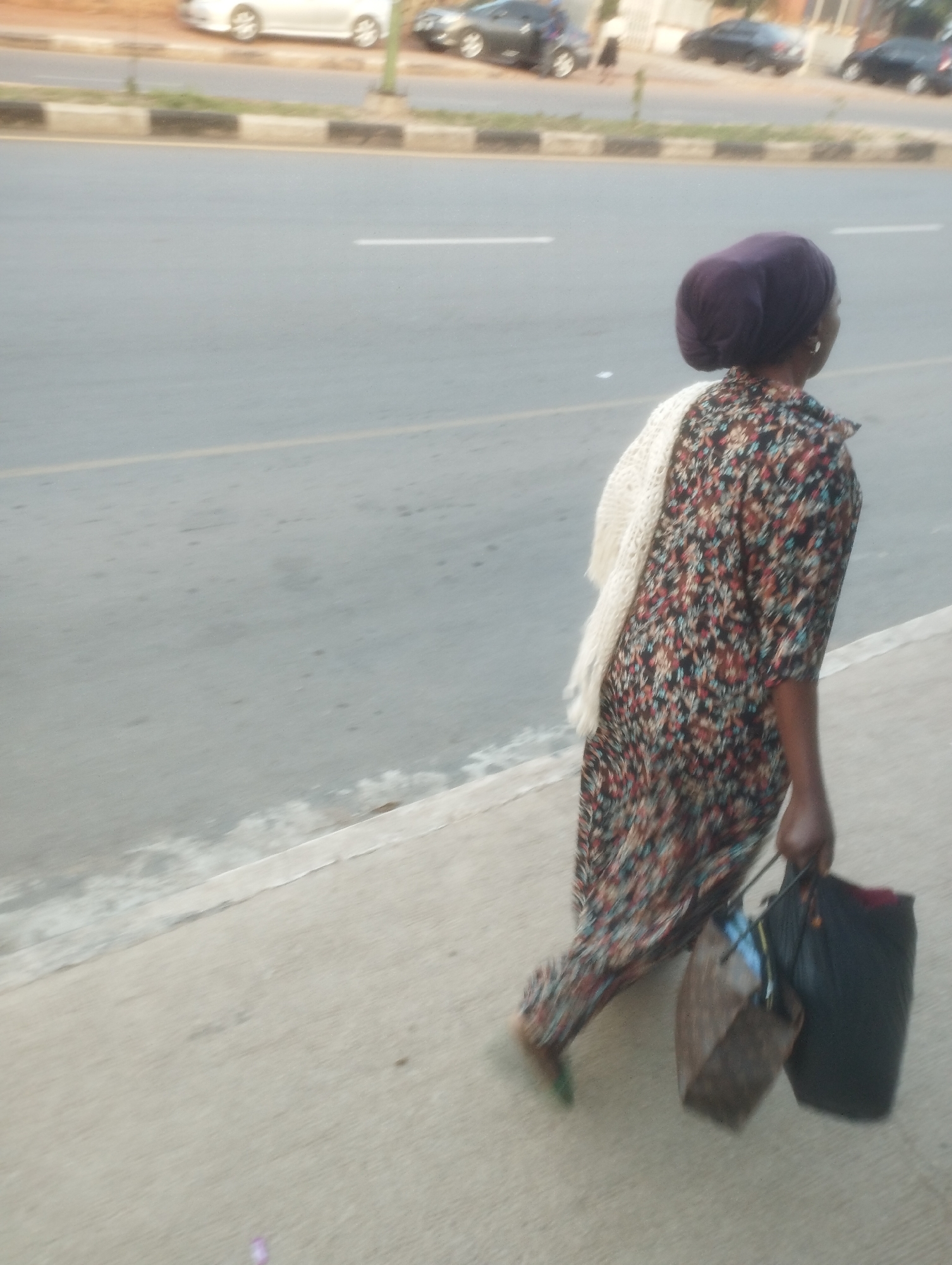 A person, viewed from behind and slightly to the side, walks along a sidewalk next to a road. The person wears a dark purple head covering, a patterned long-sleeved dress with floral motifs in shades of red, blue, and brown, and a cream-colored knitted shawl draped over their shoulders. They are carrying two bags: a dark brown, boxy bag in their left hand and a larger black bag in their right hand. Their feet are visible, wearing green footwear. The sidewalk is made of concrete, with a textured surface. Beyond the sidewalk is a paved road with a white lane marker. In the background, on the far side of the road, several vehicles are present, including a silver car, a dark-colored sedan, and another dark-colored vehicle. The overall environment suggests an urban setting. The image is consistent with a scene captured in the city of Jos, Nigeria, given the typical urban streetscape and potential for pedestrians carrying goods.