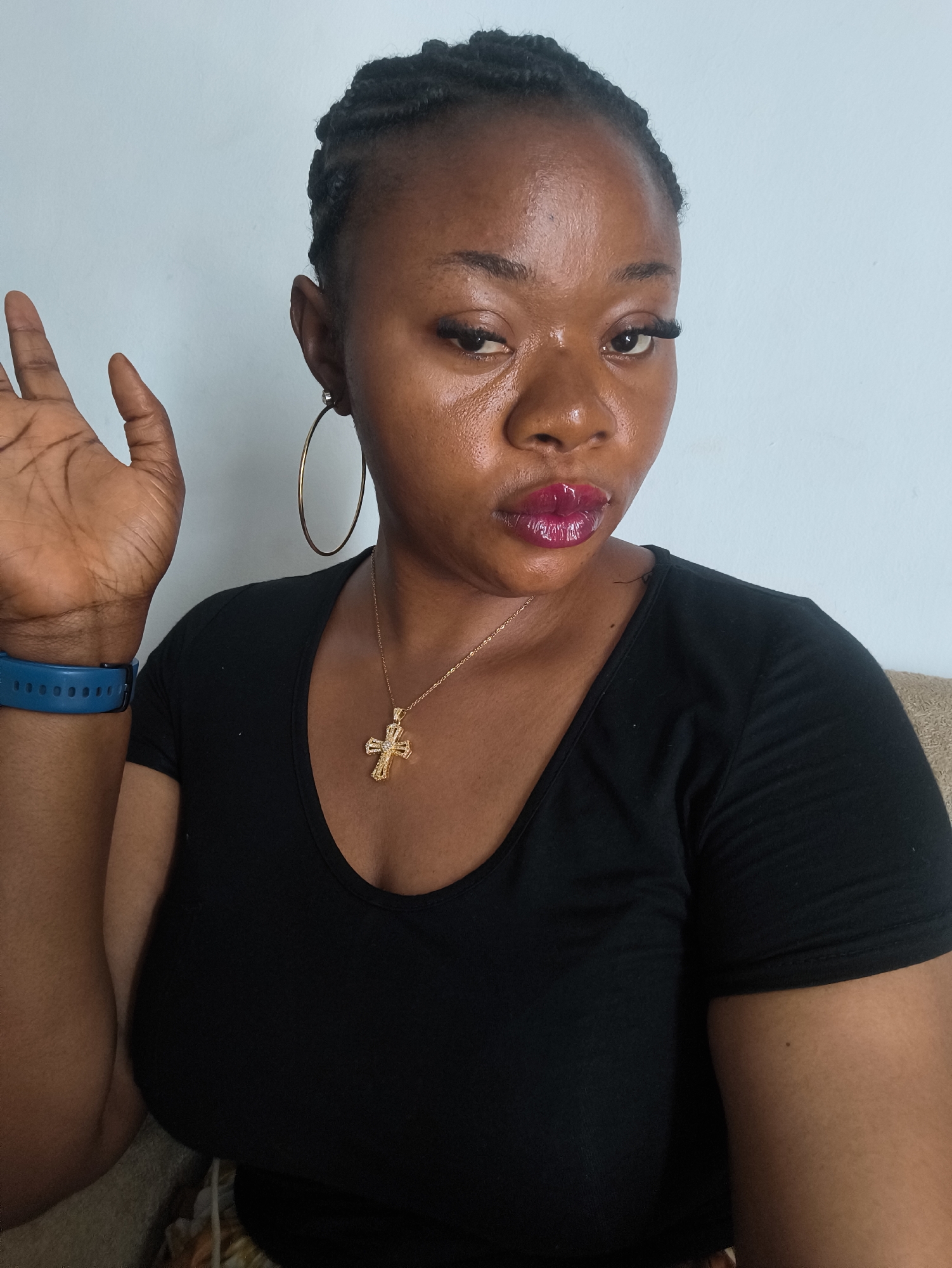 A close-up, eye-level portrait features a young Black woman in an indoor setting in Jos, Nigeria. She has dark braided hair pulled back from her face. Her complexion has a noticeable sheen on her forehead and nose. She is wearing makeup, including prominent false eyelashes and glossy, bright purple lipstick. Large gold hoop earrings are visible on both ears. Around her neck, she wears a gold chain with an ornate, jewel-encrusted gold cross pendant.
The woman is dressed in a black, short-sleeved, scoop-neck top. A blue fitness tracker or smartwatch is on her left wrist. Her left hand is raised beside her head, palm open and facing the viewer, with fingers slightly spread. She looks directly at the camera with a focused, unsmiling expression, her head slightly tilted.
The background is a plain light blue wall. A light brown or beige sofa cushion is visible in the lower right corner and along the bottom edge, suggesting she is seated. The lighting is bright and even, indicating an indoor daytime environment. No text is visible in the image.