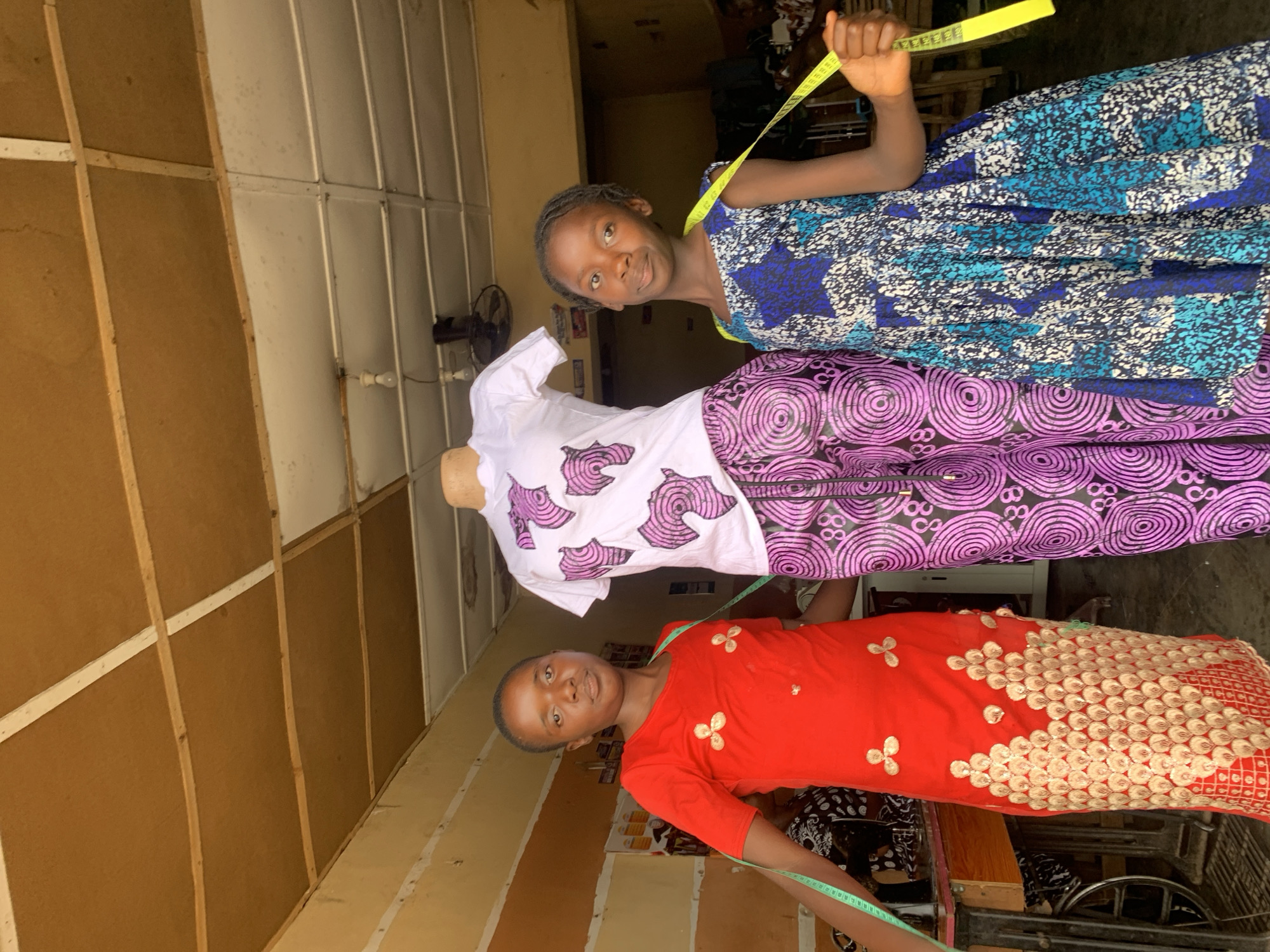 Two young girls pose with a dressmaker's dummy in what appears to be a tailoring shop in Jos, Nigeria. The girl on the right wears a red dress with gold embellishments and has a green measuring tape draped around her shoulders. The girl on the left wears a blue patterned dress and has a yellow measuring tape around her neck. The dummy is dressed in a white top with pink designs and purple patterned trousers. The setting appears to be a small, cluttered workspace with a sewing machine visible in the background. The walls are made of corrugated metal sheets.
