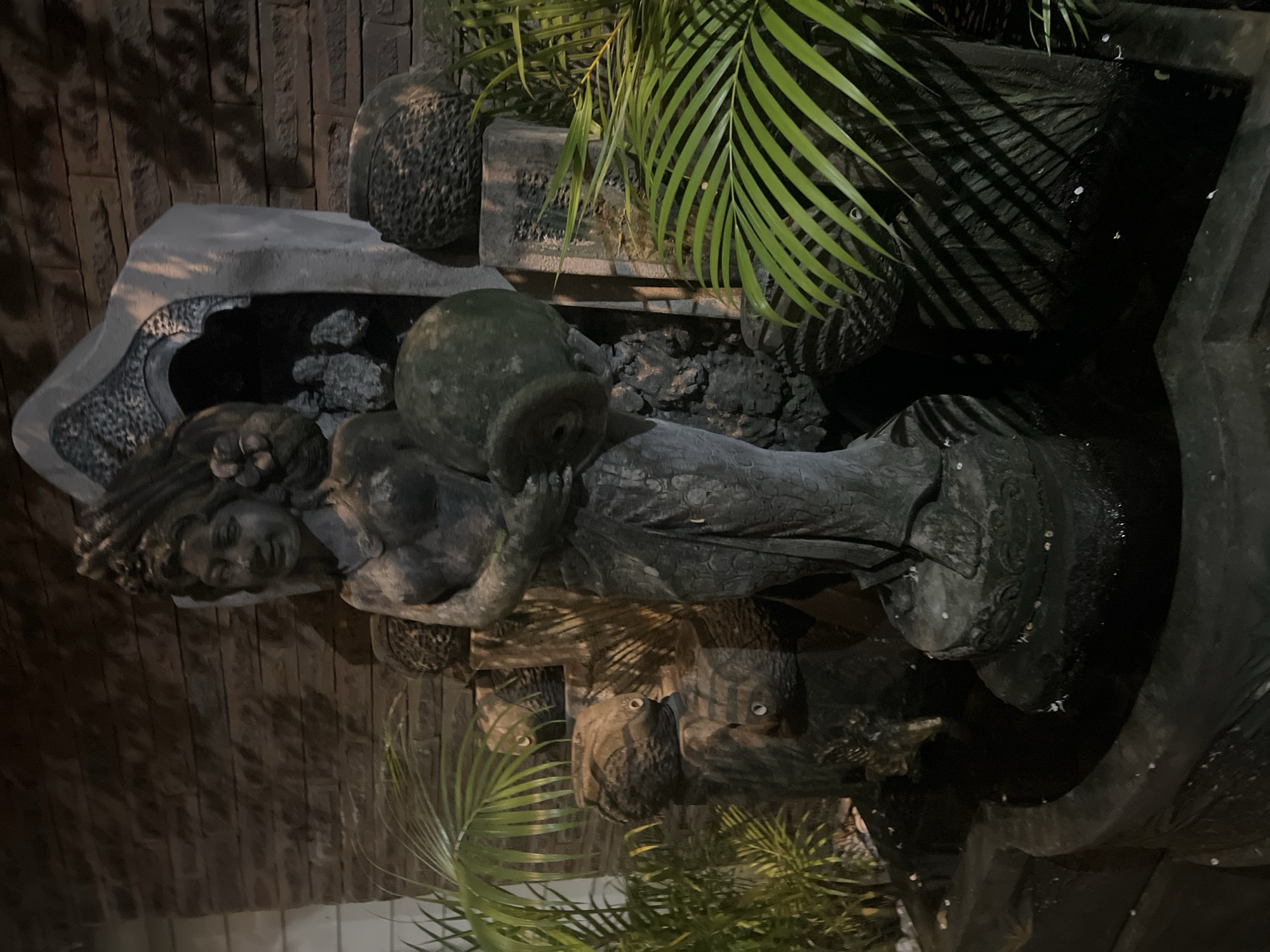 The image captures a statue of a woman, likely part of a fountain or decorative water feature, set against a backdrop of a brick or stone wall in what appears to be a garden or outdoor space at night. The main subject is a grey, weathered stone statue of a woman, depicted from the waist up, holding a large, round pot or urn. Her hair is intricately sculpted, and her face is visible, looking slightly upwards. She is adorned with what appears to be traditional or decorative attire, showing textured patterns.
The statue is integrated into a larger structure, with a basin or trough below it, suggesting it functions as a fountain. Several dark, irregular rocks or stone elements are visible within this basin. Lush green palm fronds and other foliage are prominently featured, casting long shadows across the scene, particularly on the wall and within the water feature. The lighting is dim, indicating it is nighttime, with artificial light sources illuminating parts of the statue and plants, creating strong contrasts and deep shadows.
Other decorative elements include textured stone pots or carvings positioned around the fountain structure. The overall aesthetic suggests a tranquil, ornamental garden setting. There is no visible text. The style of the statue and the lush greenery align with the provided context of North Kuta, Indonesia. No specific activity or event is taking place beyond the static display of the garden feature.
