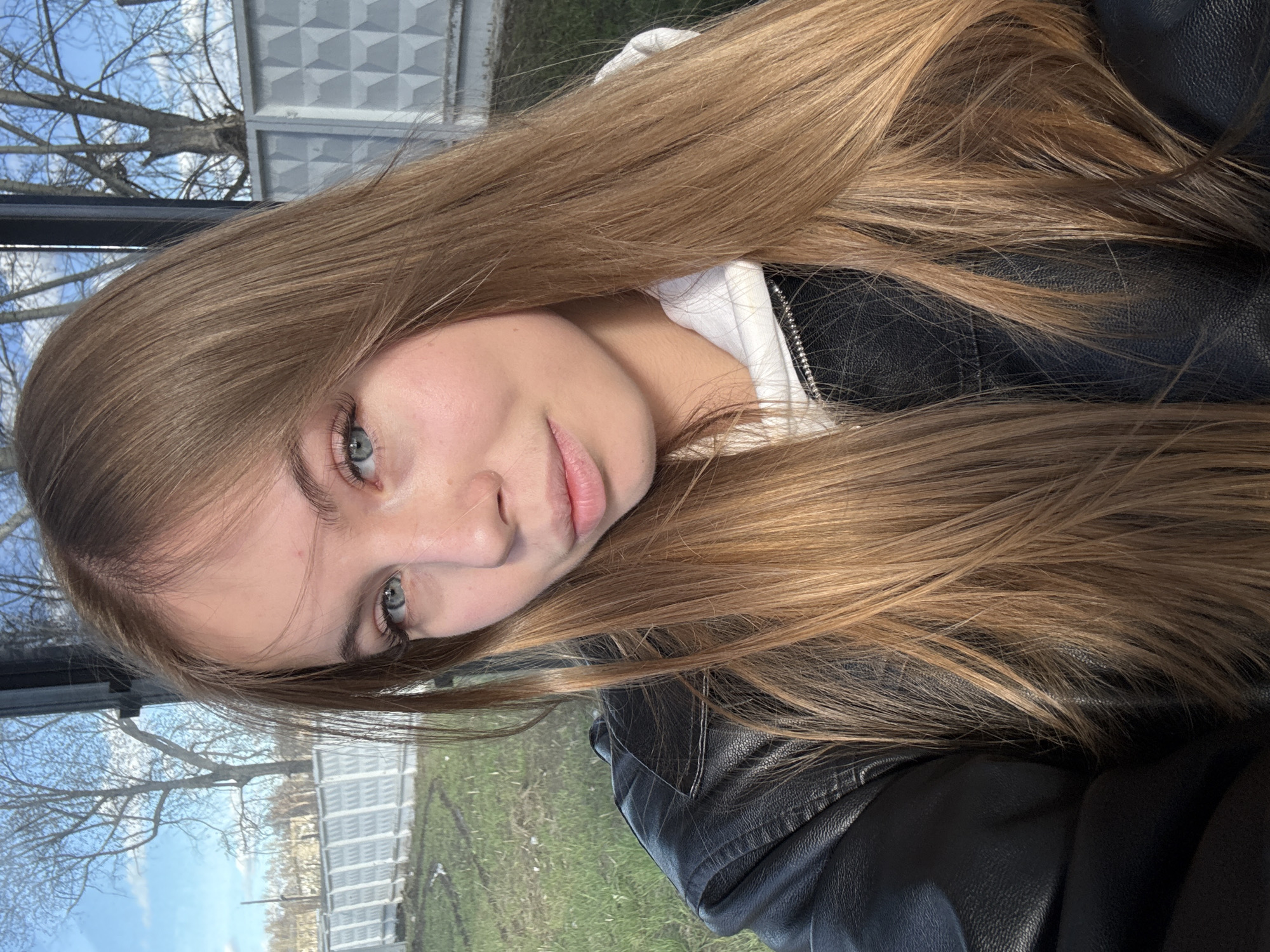 A woman is posing in an indoor setting in what appears to be Chaikovsky, Russia. She has light-brown, long hair, pale skin, and grey eyes. She wears a white hooded sweatshirt under a black leather jacket. Through the windows, there is a view of trees without leaves, a light-green lawn, and a gray building.