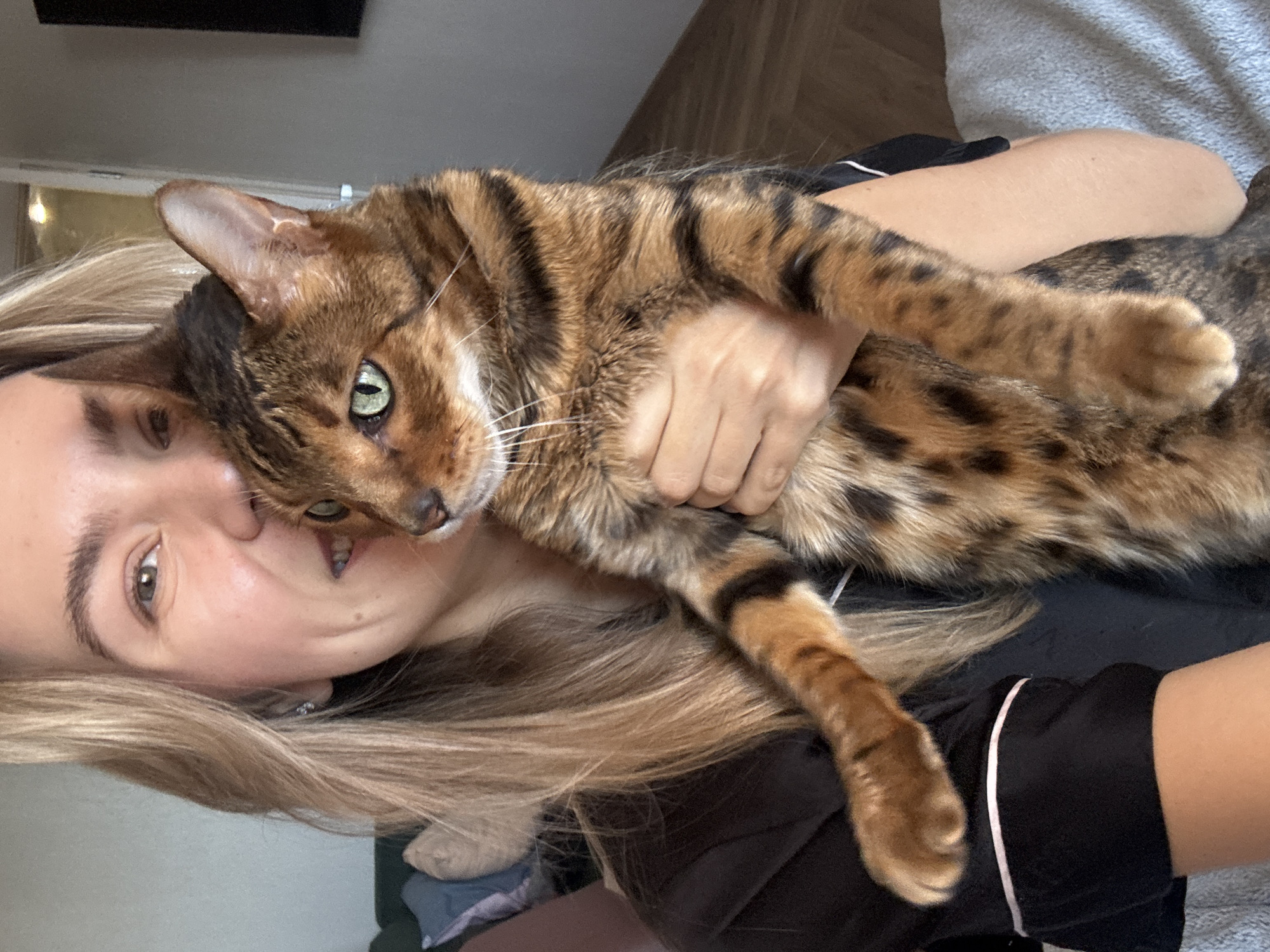 A woman is holding a bengal cat. The woman has long, blonde hair and is wearing a black shirt with white trim. The cat is brown and black with green eyes. The location is Unknown, Unknown.