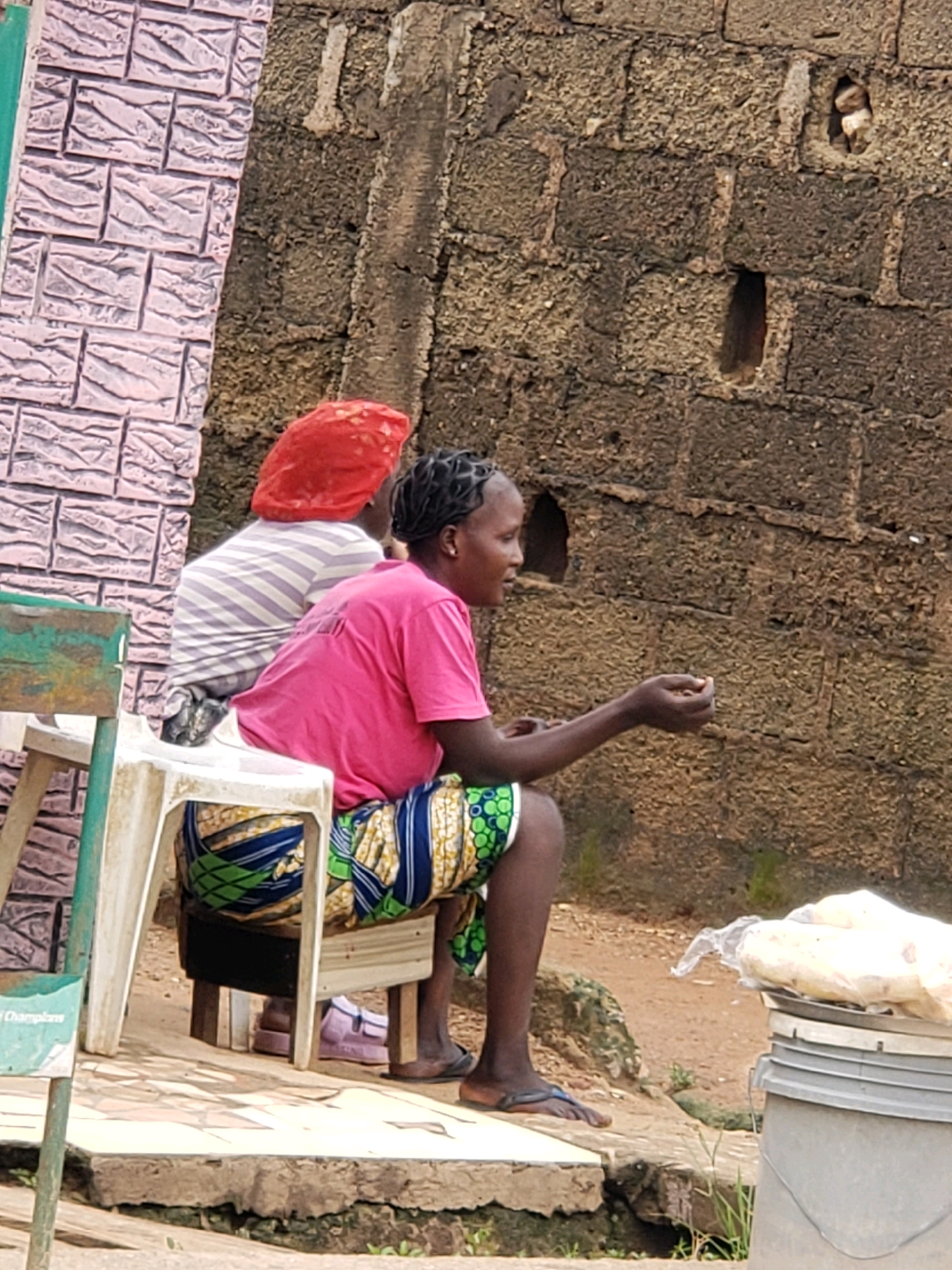 In Narayi, Nigeria, two women are seated outdoors on plastic chairs. The woman on the right, wearing a bright pink t-shirt and a patterned skirt with green, blue, and yellow motifs, is looking to her left and holding a small object in her right hand. Her braided hair is visible. To her left, a second woman, partially obscured, wears a striped shirt and a red headscarf. She is seated behind the first woman. They are positioned in front of a textured, dark brown brick wall. To the left of the scene, a pink brick wall with a textured pattern is visible, along with a portion of a green metal structure. The ground appears to be a mix of concrete and dirt. A grey bucket with white items covered in plastic wrap is visible to the right of the seated women.