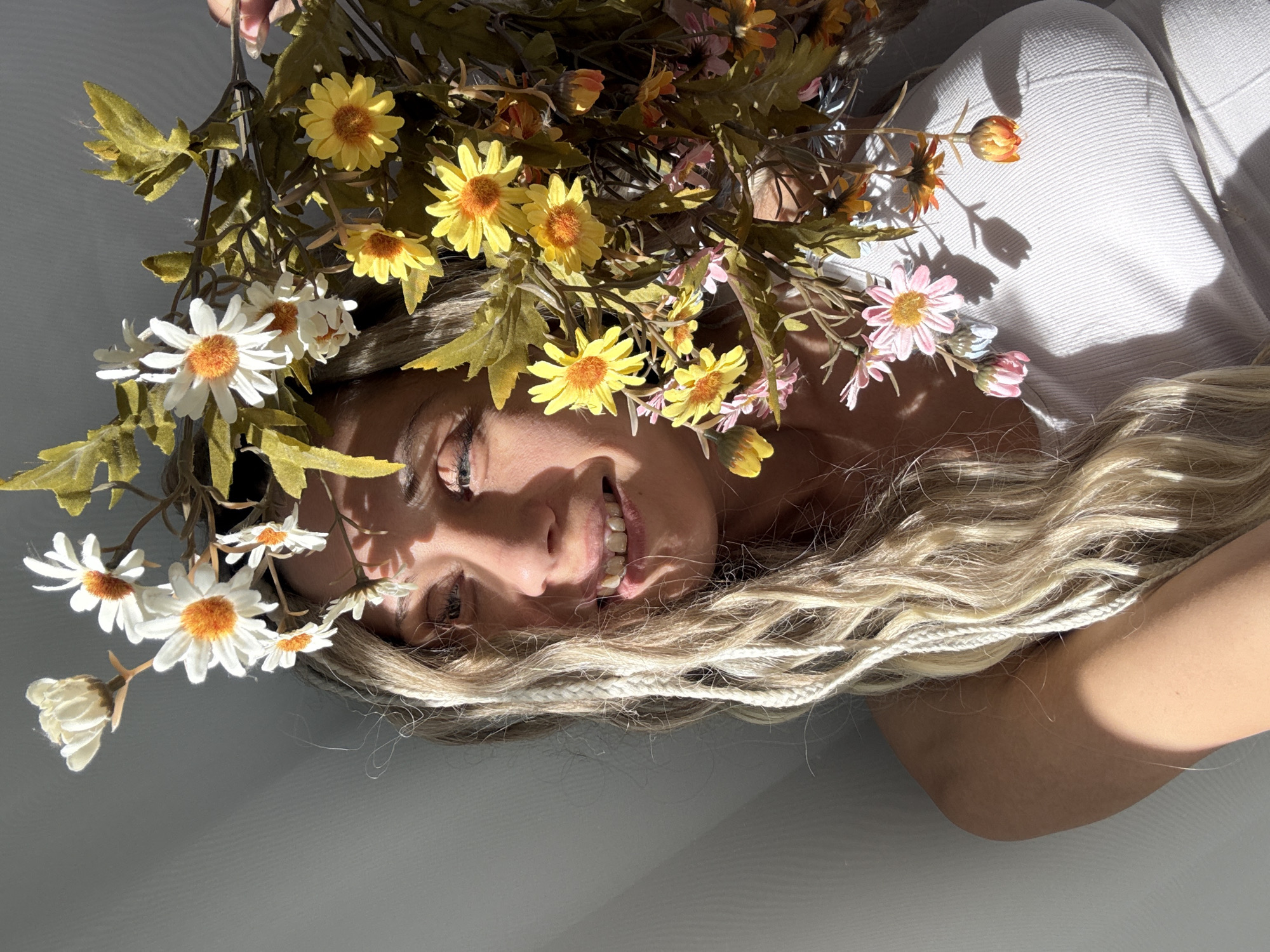 An overhead, close-up shot captures a person, likely female, lying down with a bright smile on her face, in what appears to be an indoor setting in Шебунино, Russia. Her eyes are partially closed due to the bright light, and she has long, wavy blonde hair, with several thinner braids visible among the loose strands.
Artificial flowers in shades of white, yellow, and light pink/orange, along with green foliage, are arranged over her head and hair, casting intricate shadows on her face and the white, textured surface (possibly clothing or bedding) she is resting on. The white daisies have prominent orange centers, as do the yellow flowers.
Strong, direct sunlight illuminates the scene, creating sharp, elongated shadows from the flowers and leaves on her skin and the surrounding surface, suggesting it is either morning or late afternoon on a sunny day. The person's expression is joyful and relaxed, indicated by her wide smile. The overall impression is one of natural beauty and lightheartedness. There is no visible text in the image.