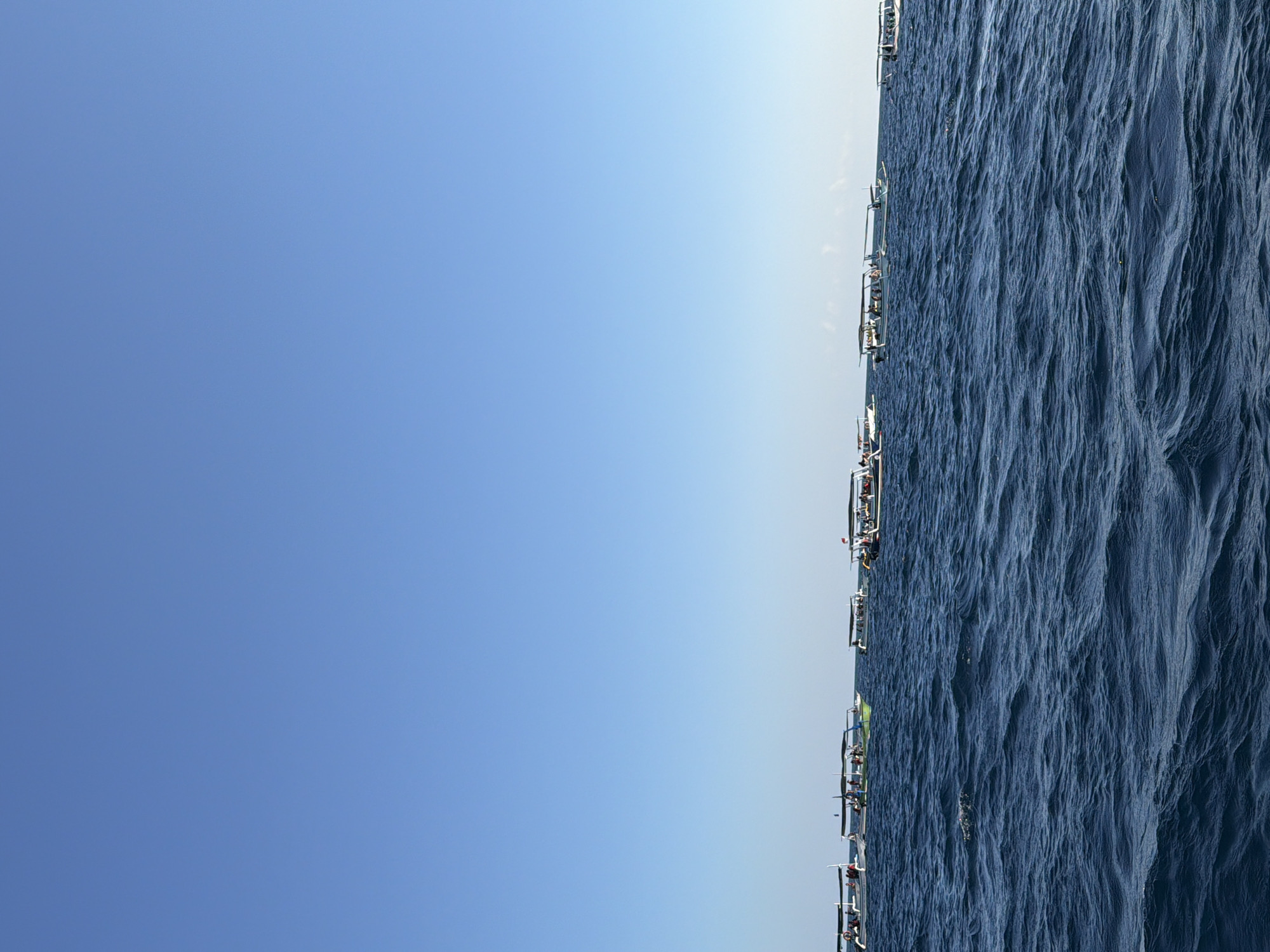 A fleet of small boats is anchored on the blue ocean under a clear sky. The boats are lined up horizontally across the image. There are people on most of the boats. The water is dark blue with small waves. The sky is a bright, cloudless blue. This scene is likely located in the city of Seririt, Indonesia, as the boats appear to be traditional Indonesian outrigger boats often used for fishing or tourism.