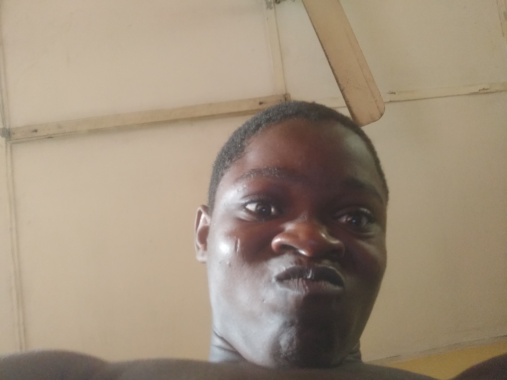 A close-up self-portrait shows a dark-skinned man with short hair, puckered lips, and a serious expression. He appears shirtless. The background includes a ceiling, a wooden beam, and part of a ceiling fan blade. The location is likely indoors. There is no indication of the city of Shangisha, Nigeria, in the image.