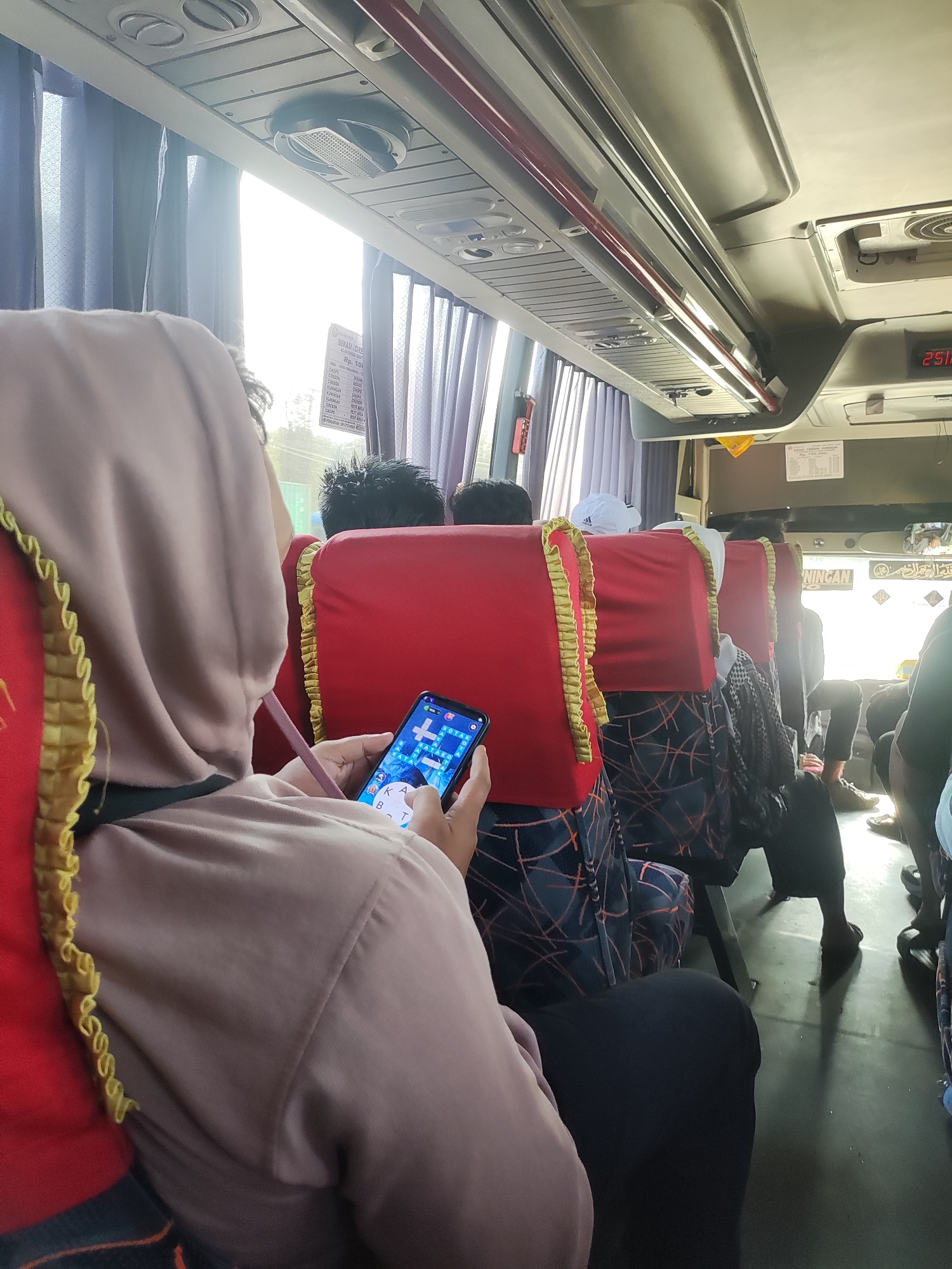 The image captures the interior of a bus in Karawang, Indonesia, as indicated by the sign that reads "BUS KARAWANG." The focus is on a passenger in the foreground, seen from the back and shoulder, wearing a light brown hijab and a dark colored top, holding a smartphone. The phone screen displays a game, possibly a word puzzle. The bus interior features red seats with decorative gold trim and patterned upholstery. Sunlight streams through the windows, illuminating the aisle. Other passengers are visible in the background, some looking towards the front of the bus, while others are obscured. The overall scene depicts a typical bus journey with passengers engaged in their own activities.