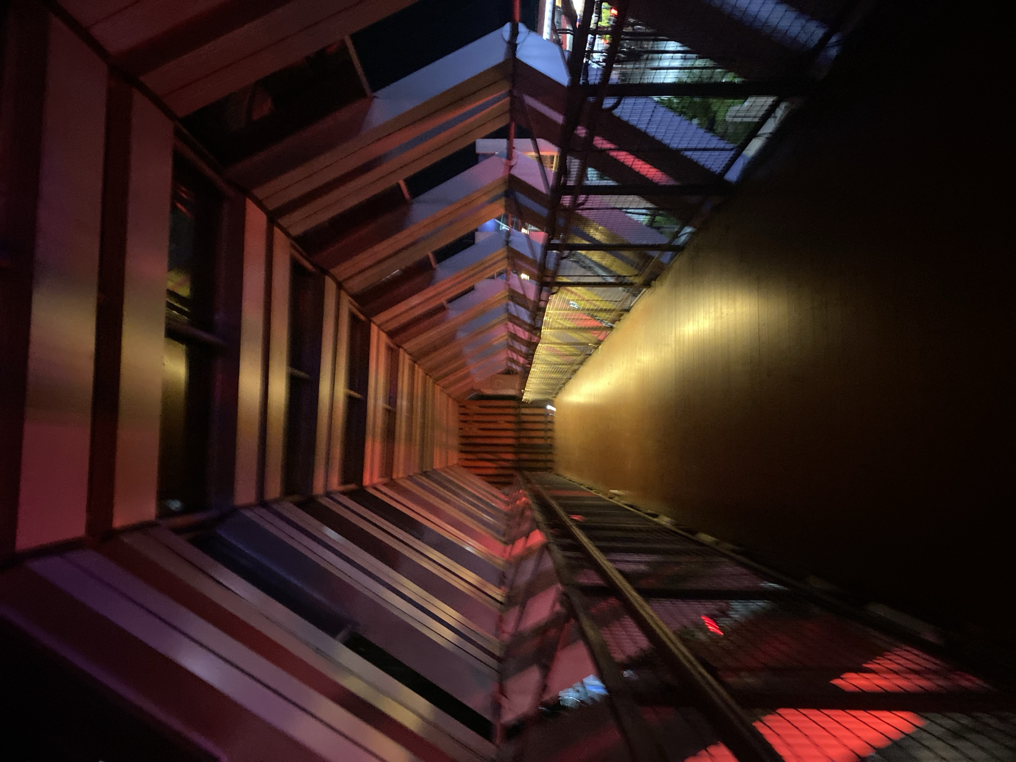 The image shows an indoor walkway with a wooden floor and metal railings. The ceiling and walls are constructed with angled metal beams, creating a tunnel-like effect. The walkway is illuminated by a warm, golden light from an unseen source, and there are red and purple lights reflecting off the surfaces. The overall atmosphere is modern and architectural. This is a pedestrian walkway located in Banjarbaru, Indonesia.