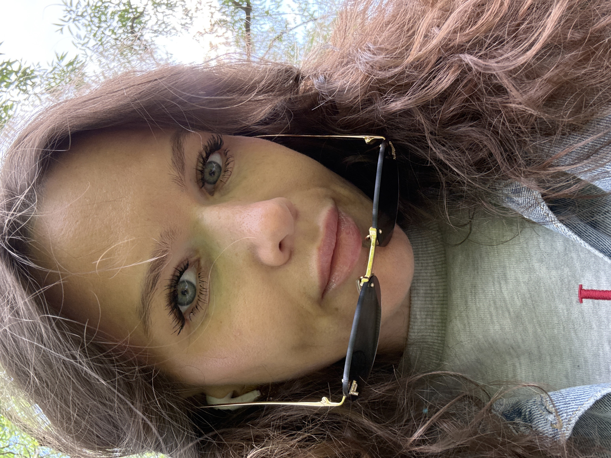 A young woman with long brown, wavy hair and green eyes is looking directly at the camera. She is wearing a light grey shirt with a small red embroidered detail on the chest and a denim jacket. A pair of sunglasses with gold frames is casually hanging on her face. A white in-ear headphone is visible in her ear. The background appears to be a park or outdoor area with greenery and sunlight filtering through the trees. It is impossible to ascertain that the location is in Moscow, Russia, from visual information.