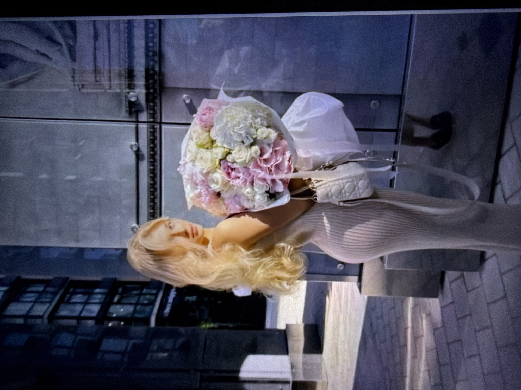 The media file shows a woman holding a bouquet of flowers and a small handbag. The woman has long, blonde hair and is wearing a light-colored, ribbed dress. The bouquet consists of various flowers, including pink roses, white hydrangeas, and possibly some pale yellow flowers. The handbag is a light-colored, quilted crossbody bag with a gold chain strap and clasp, appearing to be a designer item.
The woman is standing outdoors, with a modern building with glass windows as the backdrop. The building's facade is reflective, showing a blurred reflection of the sky and possibly other elements of the environment. In the foreground, there are paved surfaces, suggesting an urban or commercial setting. The overall lighting suggests it is daytime, possibly late afternoon, given the angle of shadows. The woman's expression appears neutral or slightly posed.
The image is oriented vertically and appears to have been taken with a mobile device. The background elements include architectural details of the building and the ground paving, which is made of rectangular bricks. There is no visible text in the image. The scene conveys a sense of casual elegance, possibly a moment captured while shopping or after receiving a gift. The location context provided is Beșalma, Moldova.