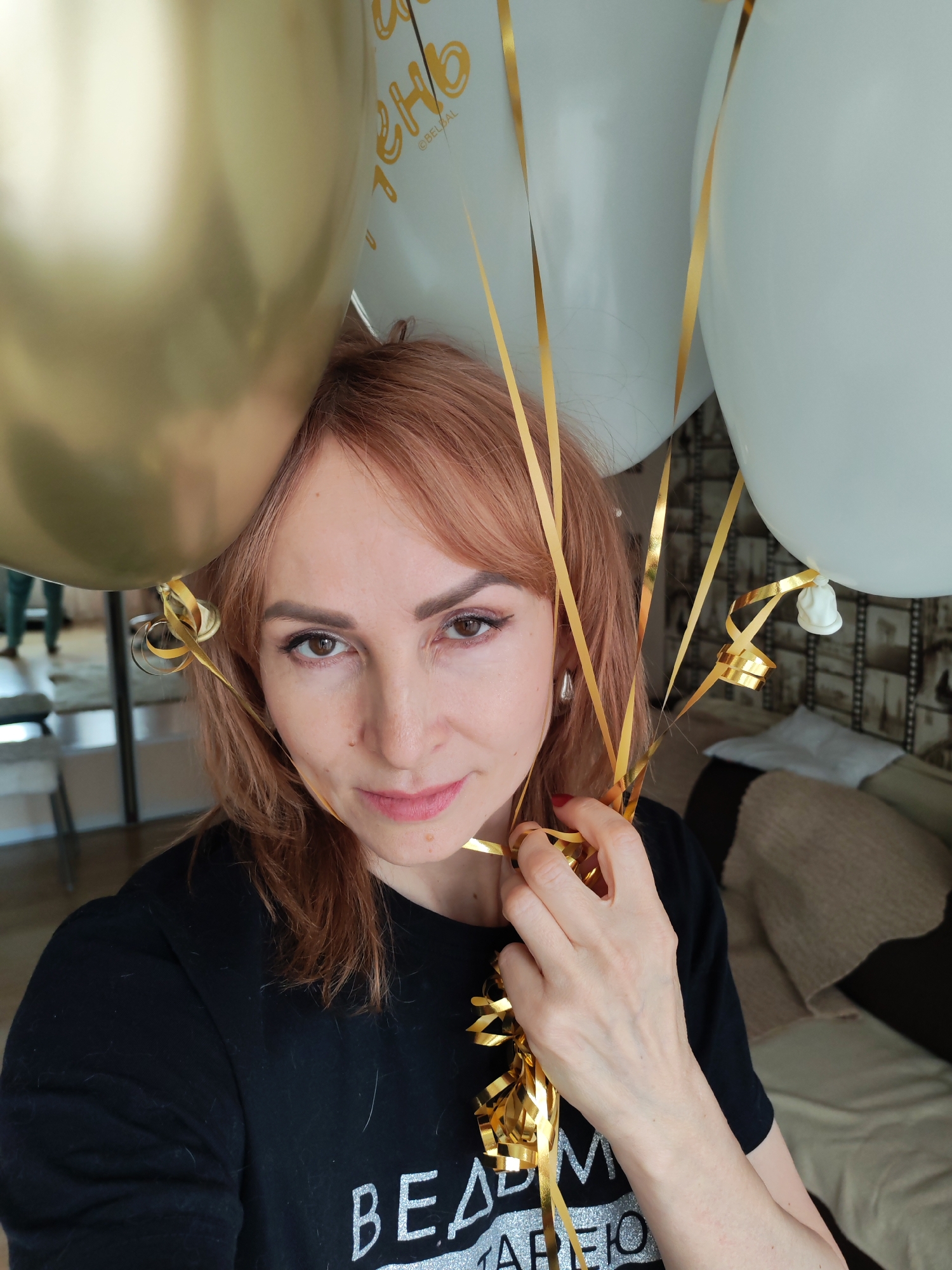 The image presents a close-up portrait of a woman in an indoor setting, likely in Chaikovsky, Russia, surrounded by celebratory balloons. She has reddish-brown hair, dark eyes, and a fair complexion, looking directly at the camera with a calm, pleasant expression and a subtle smile. A small mole is visible on her left cheek (viewer's right).
The woman is wearing a black long-sleeved top, which features silver glittery text. Part of the text reads "BEA", followed by partially visible Cyrillic letters that appear to form "ДМ Я СТАРЕЮ" (DM YA STAREYU), translating to "I AM GETTING OLD". Her right hand (viewer's left) holds a cluster of shiny gold ribbons, which are attached to a mix of golden metallic and white balloons. A large golden balloon is prominently positioned in the upper left, partially framing her. Several white or light blue balloons are also visible, one of which clearly displays the Cyrillic word "ДЕНЬ" (DEN', meaning "DAY") and the brand "OBELKAL". A silver-colored, teardrop-shaped earring is visible on her right ear (viewer's left).
The background is softly blurred, revealing parts of a home interior. A light-colored sofa or couch, partially covered by a brown textured throw blanket, is seen behind her. A patterned wallpaper or wall covering is visible further back. The overall scene suggests a festive event, possibly a birthday celebration, given the balloons and accompanying text. The lighting indicates it is daytime.