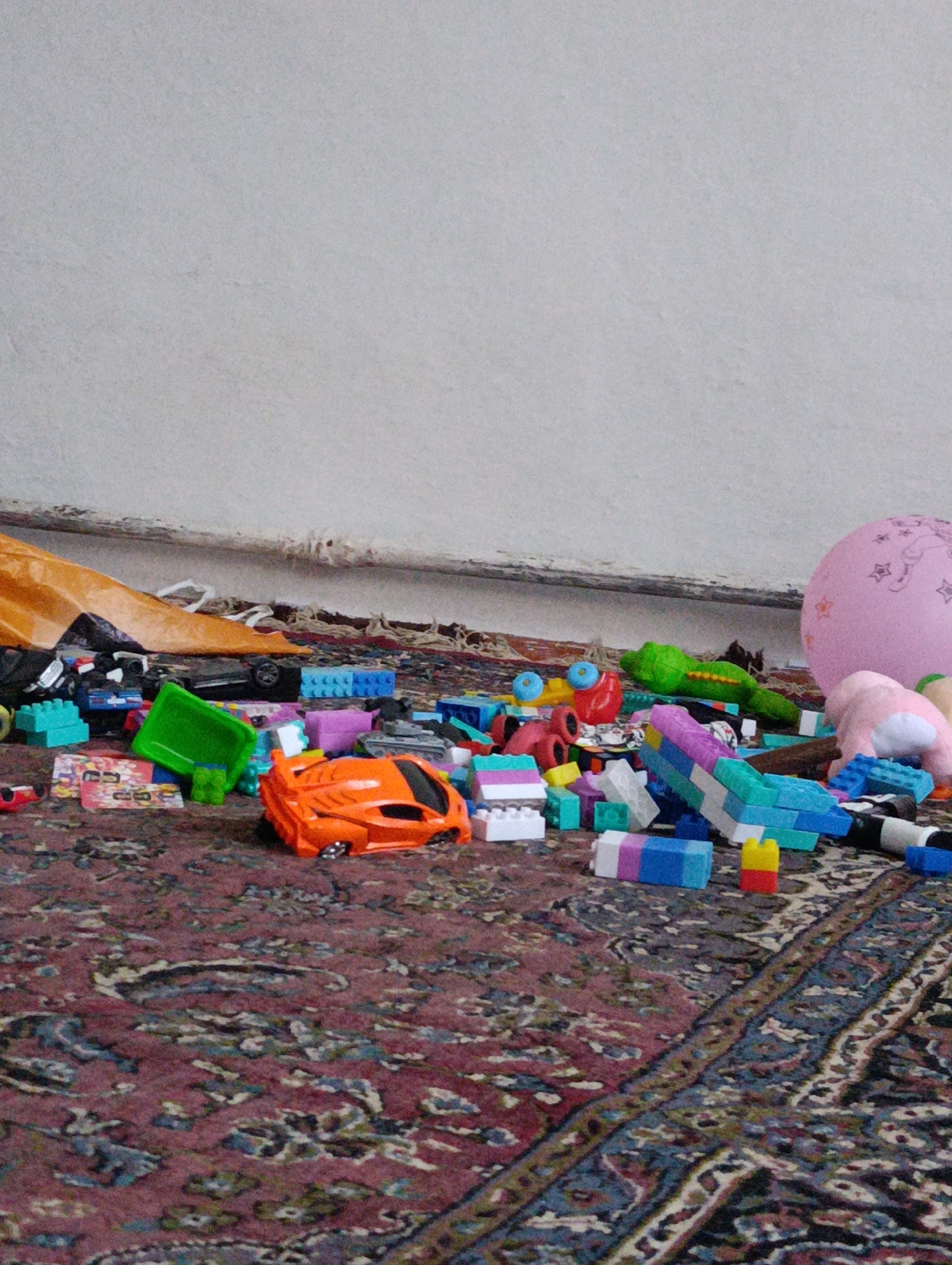 A cluttered floor, likely in a home in Novo-Voznesenovka, Kyrgyzstan, is covered with various children's toys. A large, patterned rug forms the base of the scene. Among the scattered items are colorful building blocks, toy cars, a bright orange sports car, a green plastic container, and a pink balloon with stars. A pink stuffed animal is also visible. The background consists of a plain wall and a pipe running along its base. The lighting appears to be natural, casting soft shadows on the floor.