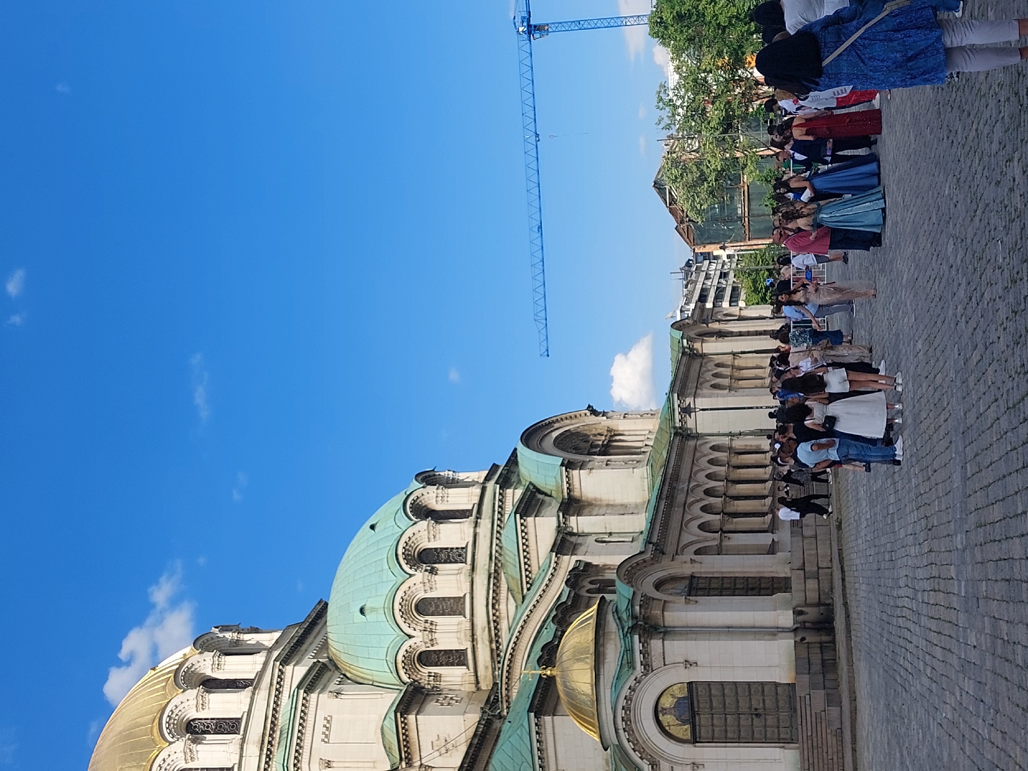 The image shows the Alexander Nevsky Cathedral in Sofia, Bulgaria. A group of formally dressed people are standing on the cobblestone plaza in front of the cathedral. A tall blue construction crane is visible in the background. The sky is clear and blue with some clouds. The cathedral is built of white stone with a green dome.