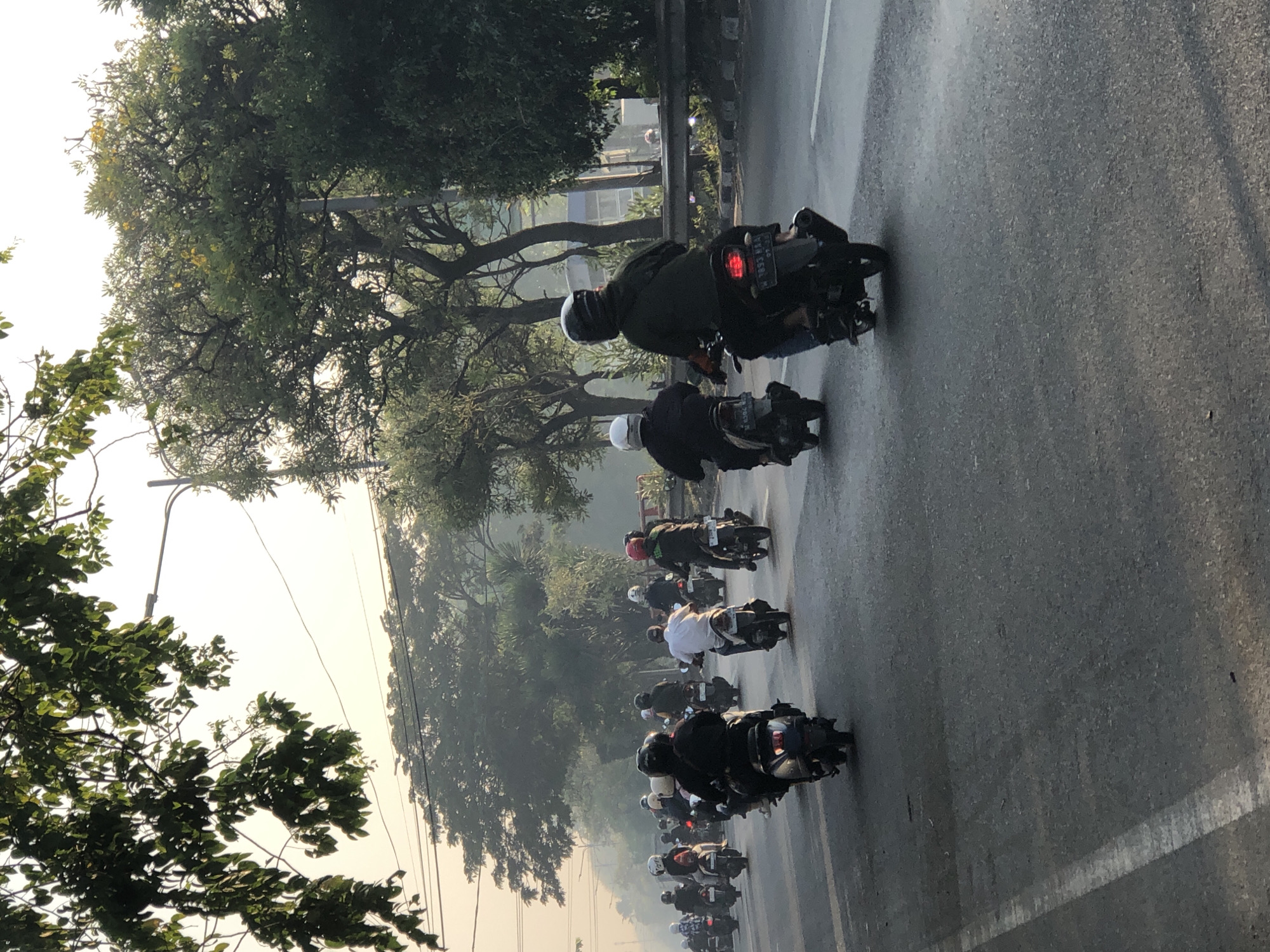A long line of motorcyclists ride on a paved road in Kludan, Indonesia. The motorcyclists are wearing helmets and riding in the same direction. Some of the motorcycles are carrying passengers. The road is lined with trees and there is a fence on the side of the road. The sky is hazy and the sun is setting. The scene appears to be a typical commute during rush hour.