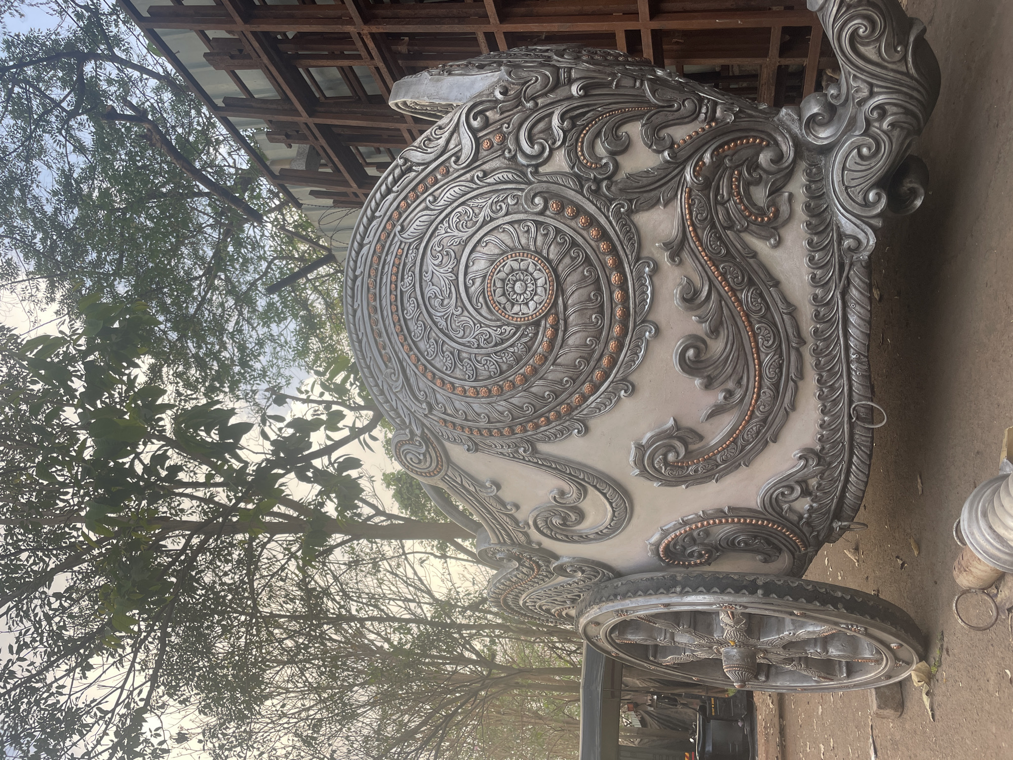 A decorative vehicle, resembling a chariot, is positioned outdoors in what appears to be a construction or storage area. The chariot is predominantly white with ornate silver and copper-toned embellishments. A large circular motif adorns the side, featuring a swirling pattern and decorative raised details. The chariot has wheels with an intricate spoke design.
The background includes a metal structure, possibly a scaffolding or framework, and trees with bare branches and sparse foliage. The ground appears to be dirt or gravel. The image is underexposed, creating a somewhat dim lighting.
While the image provides visual details, the precise location within Mumbai, India, cannot be determined without additional information or context.
