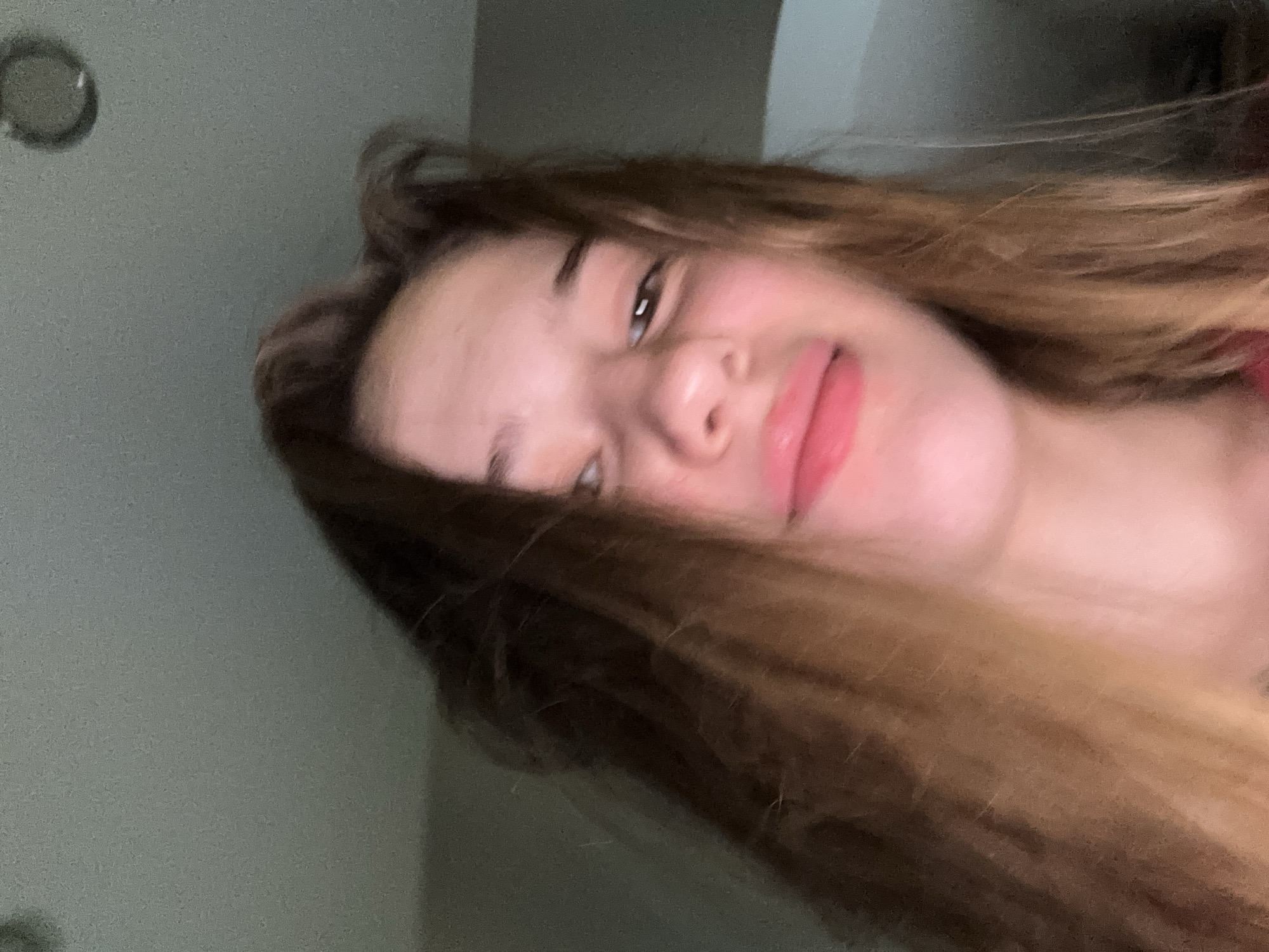 A young girl with long, light brown hair is looking at the camera with a slight smile. Her lips are slightly parted and appear to be wearing lip gloss. The lighting is dim, casting a shadow on her face. The background is out of focus, but appears to be an indoor setting with a ceiling light visible. There is no discernible location in the city of Unknown, Russia.