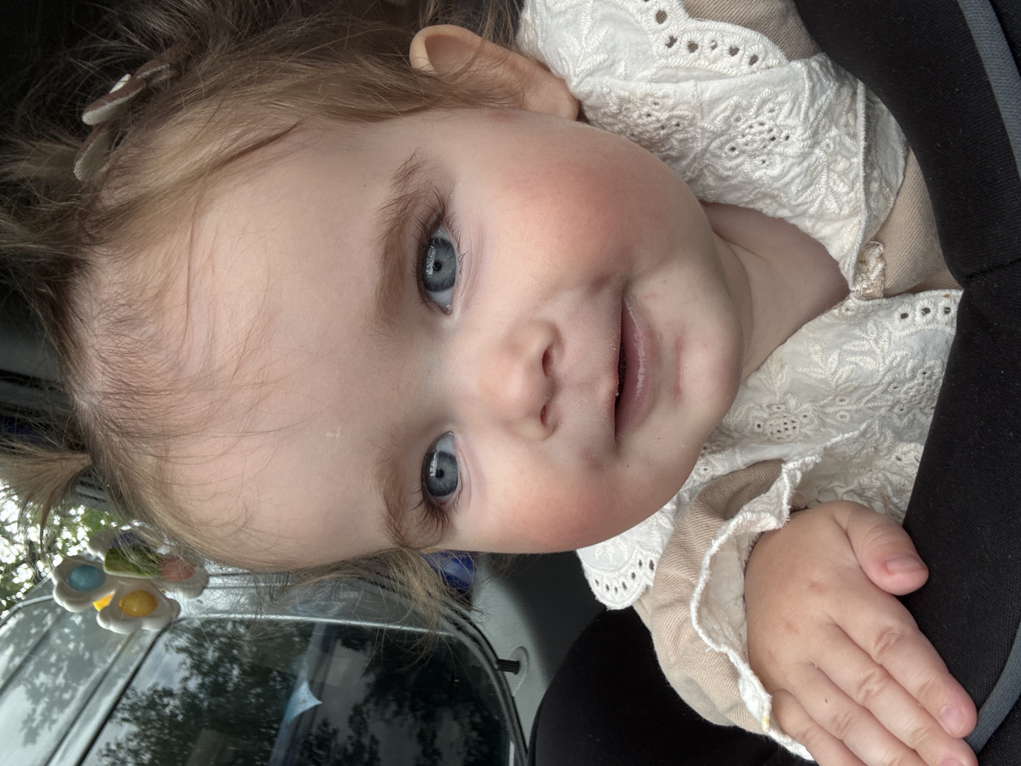 A close-up, eye-level portrait captures a fair-skinned baby girl from the chest up, looking forward with a gentle smile. She has fair skin, plump cheeks with a rosy hue, bright blue eyes, and light brown hair pulled into two pigtails, adorned with small hair clips. Her expression is content and alert. The baby is wearing a light-colored garment with intricate white lace detailing around the collar and sleeves. Her left hand rests on a black surface, possibly a car seat or stroller. In the background, out of focus, reflections of trees and foliage are visible on a dark, reflective surface, suggesting an outdoor environment. The overall lighting is soft and natural, highlighting the baby's features. The image is likely taken in Ulmen, Germany, given the surrounding environment, but no specific landmark or definitive indicator is present within the frame itself.