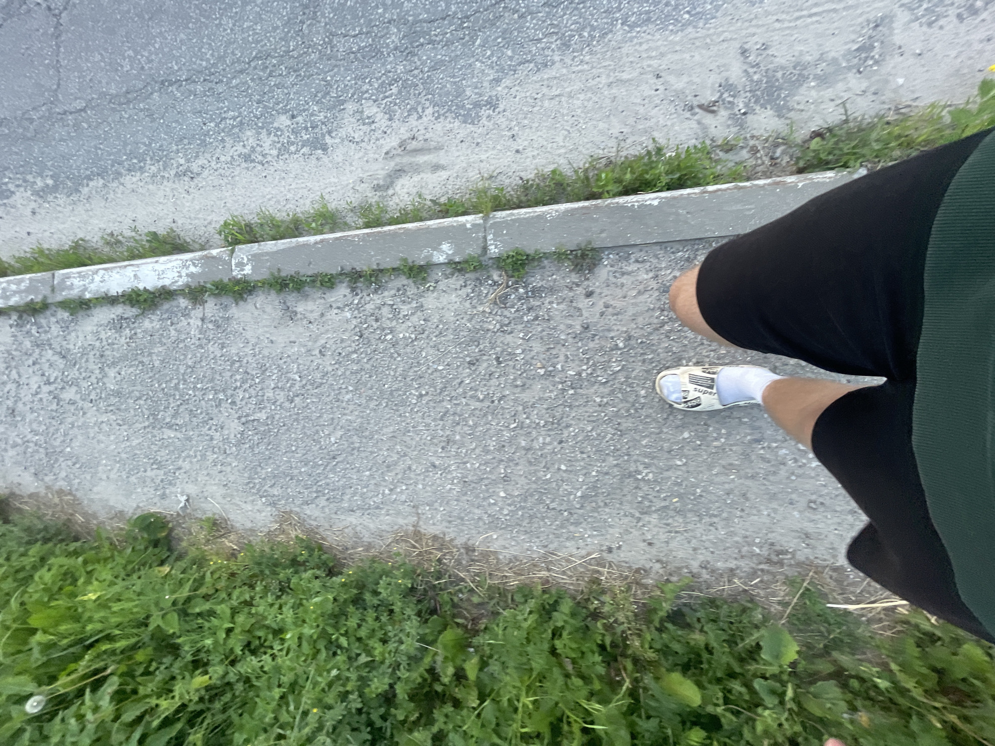 An overhead perspective shot captures a person's legs and feet as they walk along a paved sidewalk. The sidewalk, composed of gravel and concrete, is bordered on one side by a strip of green grass and vegetation. On the other side, the sidewalk meets a road that appears to be asphalt. The person is wearing black shorts, white socks, and white shoes with a black and white pattern. The environment suggests an outdoor setting, possibly in a park or along a street in the town of Krasnye Baki, Russia. The lighting indicates it's daytime, with soft shadows suggesting it may be late afternoon or early morning.