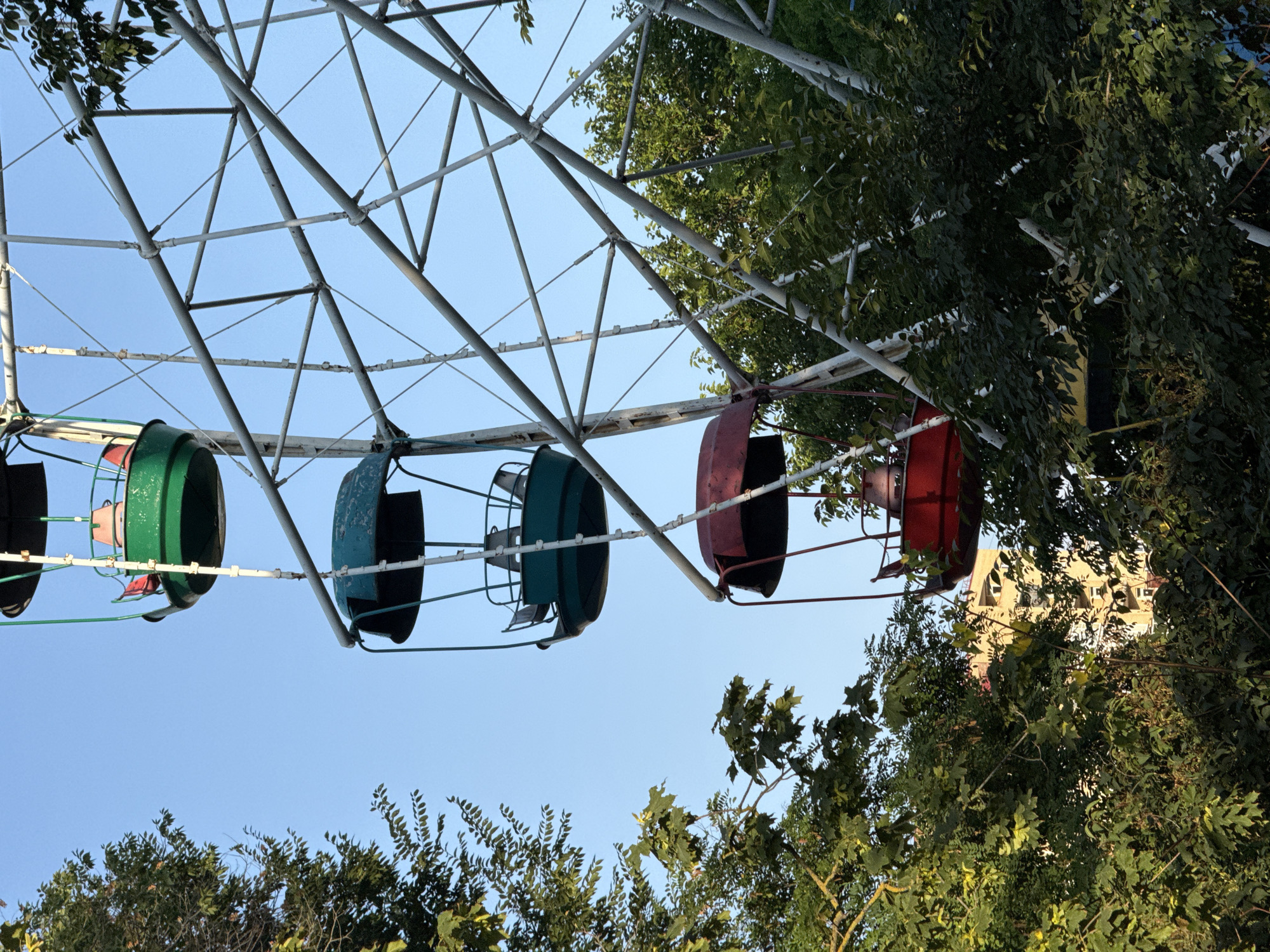 An outdoor, eye-level, medium shot captures a segment of an amusement park Ferris wheel against a clear blue sky. The Ferris wheel structure is a lattice of white and grey metal beams and diagonal supports, some of which are visibly rusted. Hanging from the wheel are four passenger gondolas, each with a red seat. The top gondola is green, followed by two teal gondolas, and then a red gondola at the bottom. Green trees with dense foliage flank the Ferris wheel on the left and right sides, partially obscuring the view. In the background, peeking through the trees and behind the gondolas, is a building with a light yellow facade and several visible windows, suggesting an urban or suburban setting. The image appears to be a photograph taken during daylight hours.