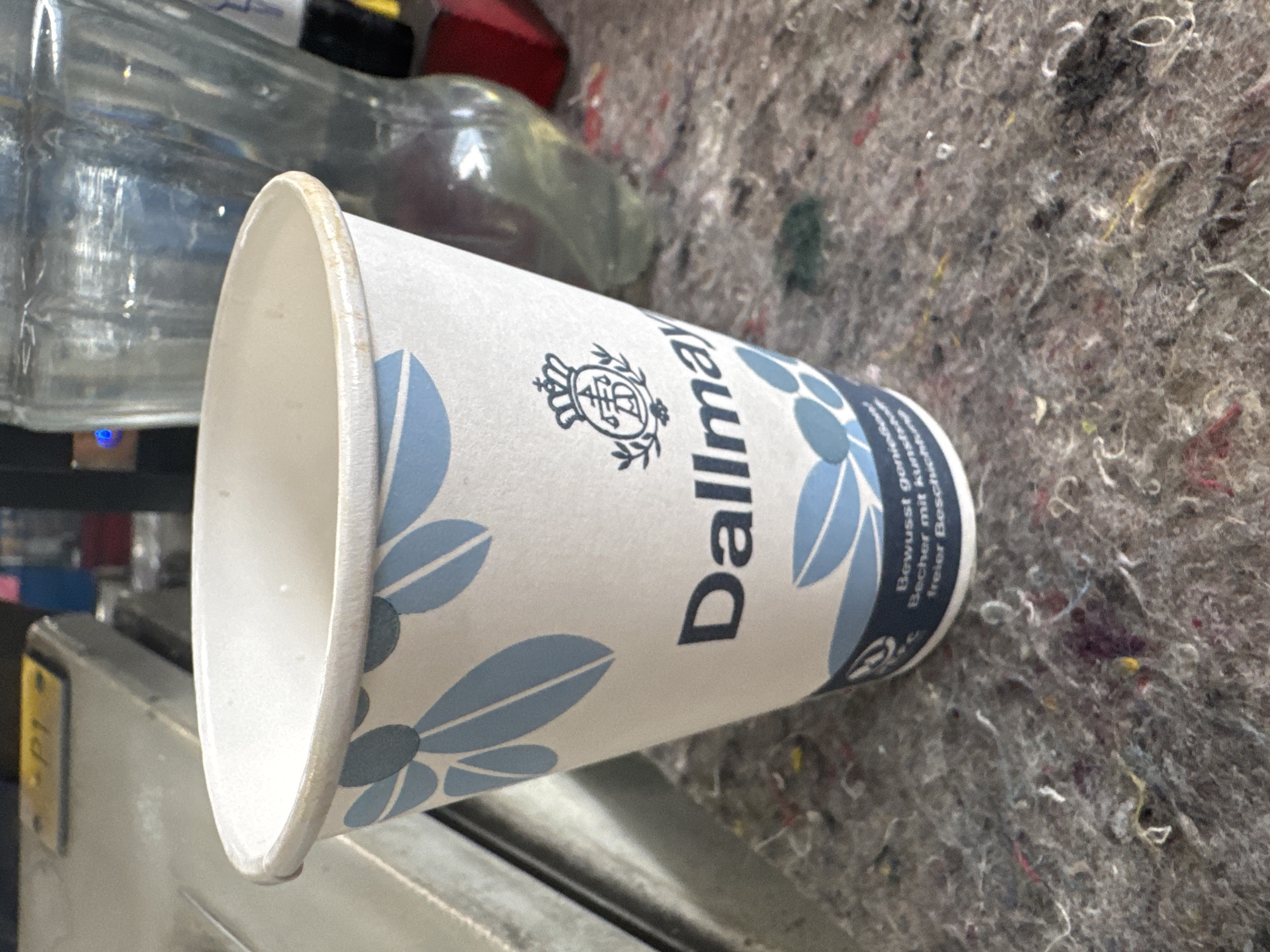 This media file captures a close-up, rotated view of a disposable paper coffee cup with a blue floral design. The cup appears to be resting on a textured, grey surface, possibly a felt or fabric material, with scattered colorful fibers. Behind the cup, blurry elements suggest a background setting, including what looks like a glass bottle and a red object.
The cup itself is white with light blue floral patterns. Prominently displayed on the cup is the "Dallmayr" logo, a circular emblem with a crown and intricate detailing, followed by the brand name in a dark, sans-serif font. Below the brand name, there is text in German: "Bewusst genießen Becher mit kur freier Beschr," which translates to "Consciously enjoy cup with short free besch." The "E" in the Dallmayr logo is also visible.
The overall lighting suggests an indoor environment. The angle of the photograph and the focus on the cup imply that the cup is the primary subject of interest, with the background providing context. No people are visible in the frame. The scene is static, with no discernible activity taking place. The location context provided is Ellwangen, Germany, though no specific visual cues within the image directly confirm this.