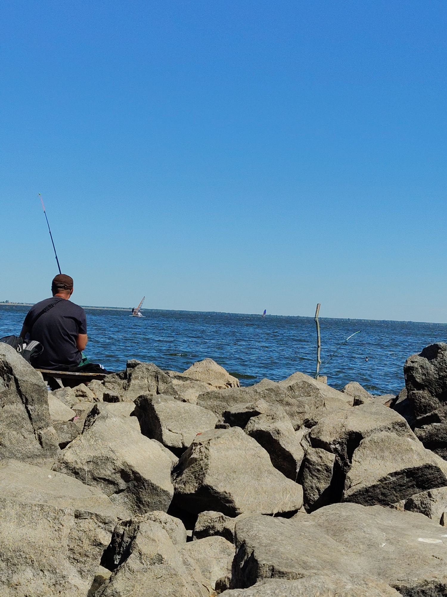 This image shows a man fishing on a rocky shore with a clear blue sky. The man is wearing a dark t-shirt and a brown cap and is seated on a wooden bench. A fishing rod is visible beside him. In the background, the blue water of a large body of water is visible, with two windsurfers in the distance. The shore is made up of large, grey rocks. The overall environment suggests a sunny day at a waterfront location. The location is identified as Cherkasy, Ukraine.