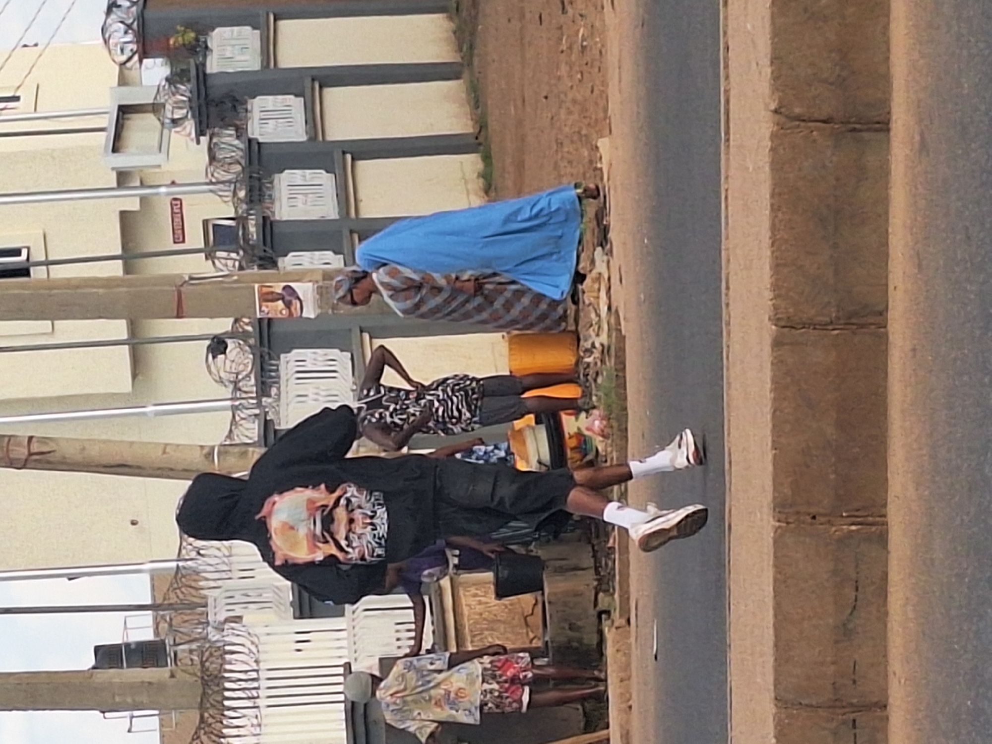 The image depicts a street scene in Sabon Tasha, Nigeria. There are several people visible, including individuals wearing casual clothing and traditional attire. Some appear to be carrying buckets and interacting near a roadside, possibly fetching water. The environment includes buildings with visible air conditioning units and security measures, along with utility poles and a paved road with a concrete barrier. The scene suggests a typical day with people engaged in daily activities.