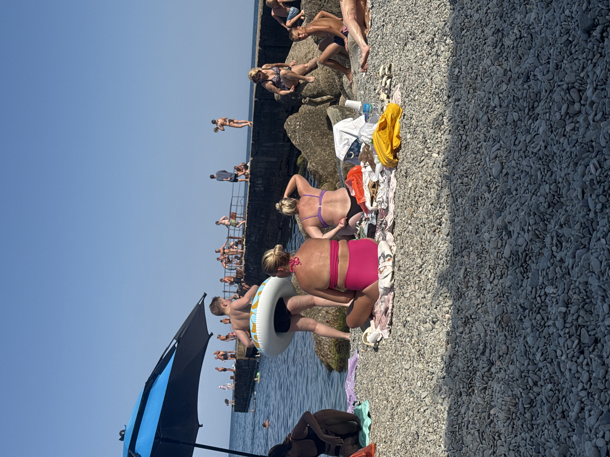 The image depicts a crowded beach scene in Partenit, Ukraine, on a sunny day. The foreground is dominated by a pebble beach, with several people sunbathing on towels. In the middle ground, a group of individuals are seated and standing on rocks near the water's edge, some in swimwear. The background shows a pier with more people, and the clear blue sea stretches to the horizon under a bright sky. An umbrella shades a portion of the beach in the left foreground.