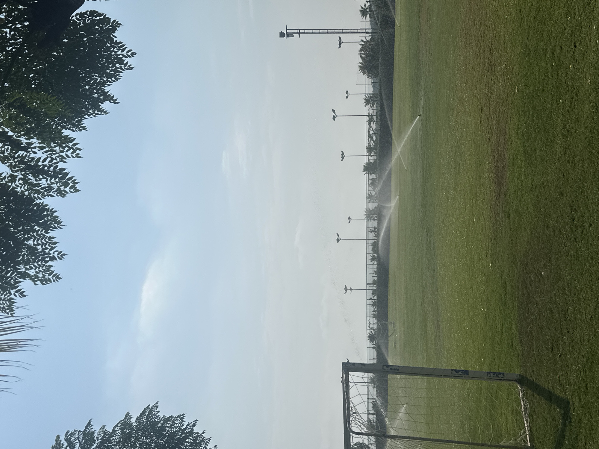 A football pitch in Manavgat, Turkey, is being watered by sprinklers. The pitch is green and well-maintained. A goalpost is visible in the foreground on the left. The sky is hazy and light blue, with some clouds. Trees and palm trees line the background, and lampposts are spaced out across the pitch. The scene appears to be early morning or late afternoon, as suggested by the soft light and haze.