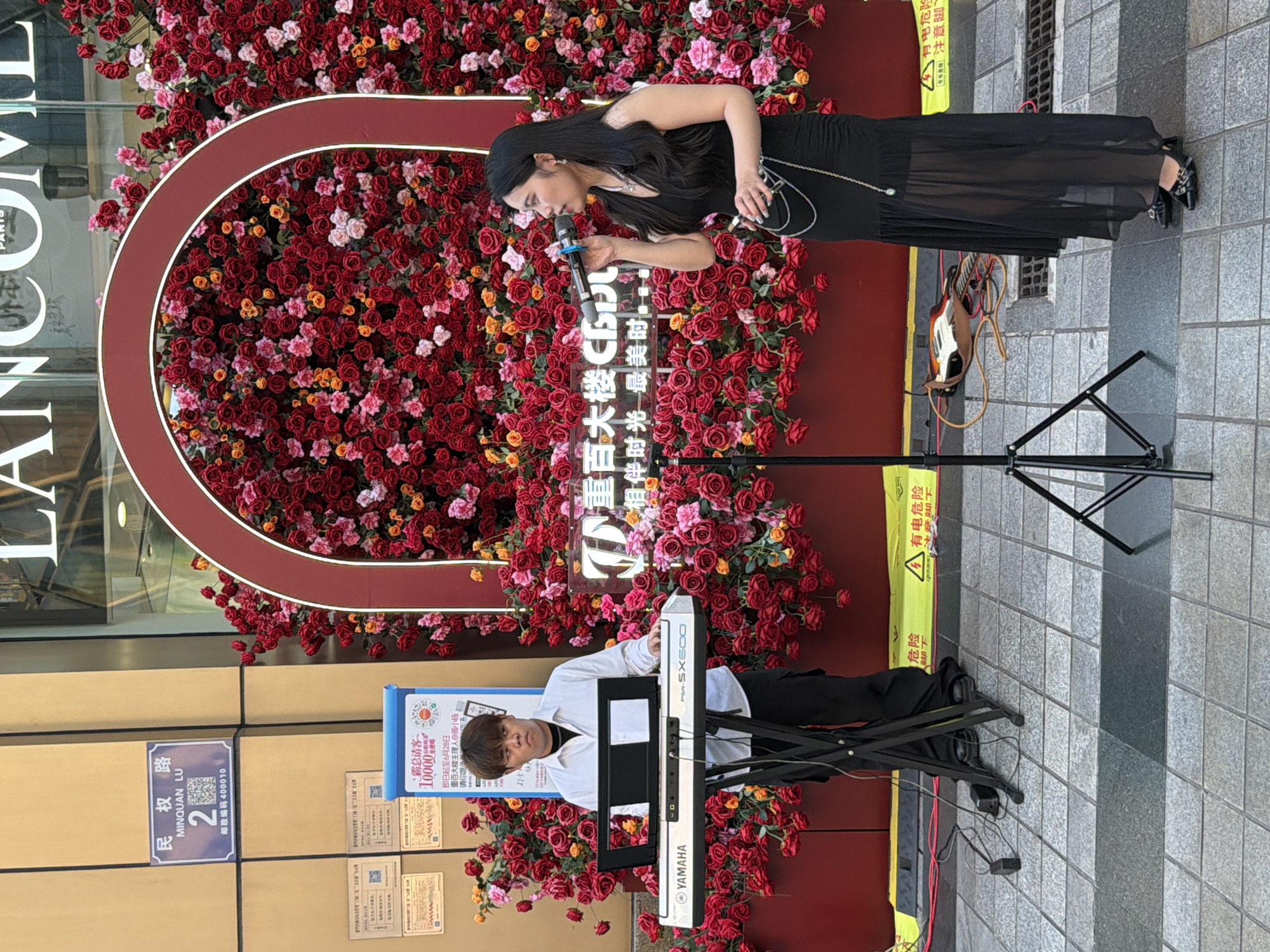 An outdoor scene in Yuzhong District, China, features two performers in front of a large floral display. A young man, wearing a white button-down shirt and black pants, is seated and playing a white Yamaha keyboard. To his right, a young woman in a black sleeveless dress and sheer skirt stands and sings into a microphone. Behind them, a wall is decorated with a vibrant arrangement of red and pink roses, framed by a burgundy archway. A neon sign with Chinese characters is visible on the wall. The ground is paved with grey tiles. Yellow caution tape with Chinese text indicating "Danger" is laid out on the ground. A guitar lies on the ground to the right of the performers. A street sign with "MINQUAN LU" and "2" is mounted on a building to the left.