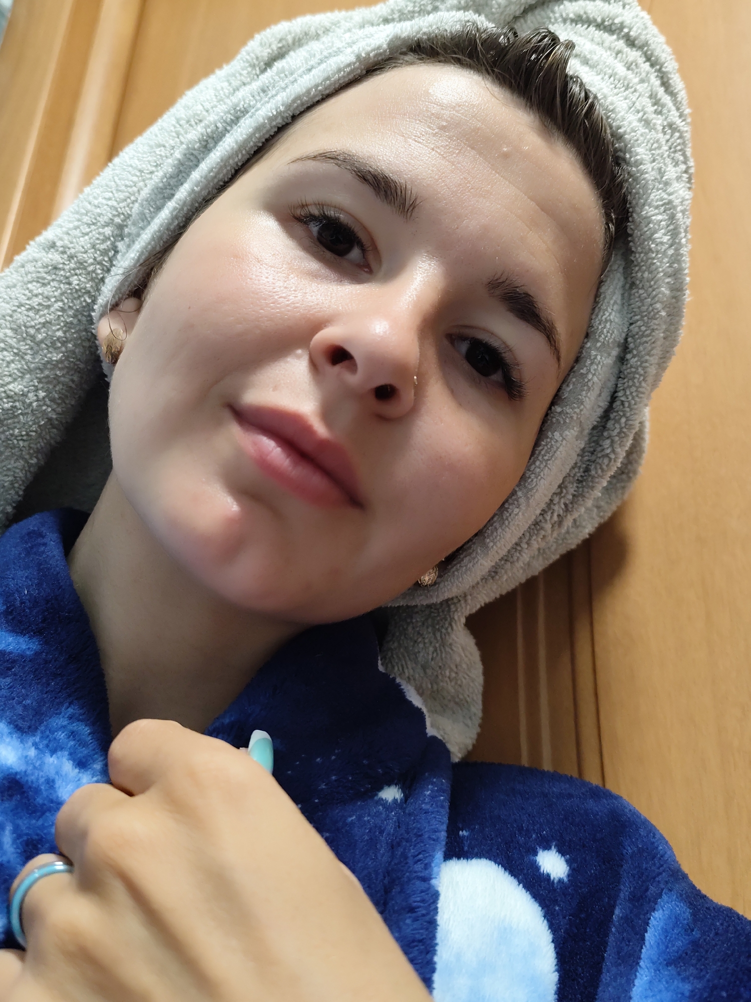 A woman with a towel wrapped around her head is indoors. She is wearing a blue bathrobe and a blue ring on her finger. A wooden door is behind her. The location is unknown.