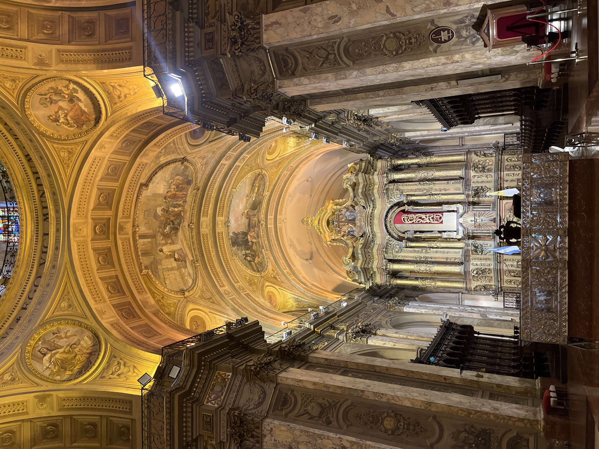 This is an interior view of the Metropolitan Cathedral of Buenos Aires, Argentina, looking up towards the dome and altar. The architectural style is predominantly neoclassical, featuring elaborate ornamentation, gilded details, and painted frescoes on the dome's interior. The dome is adorned with classical imagery, including figures and decorative motifs. Massive columns and detailed stonework frame the view of the altar area, which is also richly decorated with gold accents and religious iconography. The image is taken from an angle looking upward, emphasizing the scale and grandeur of the cathedral's interior.