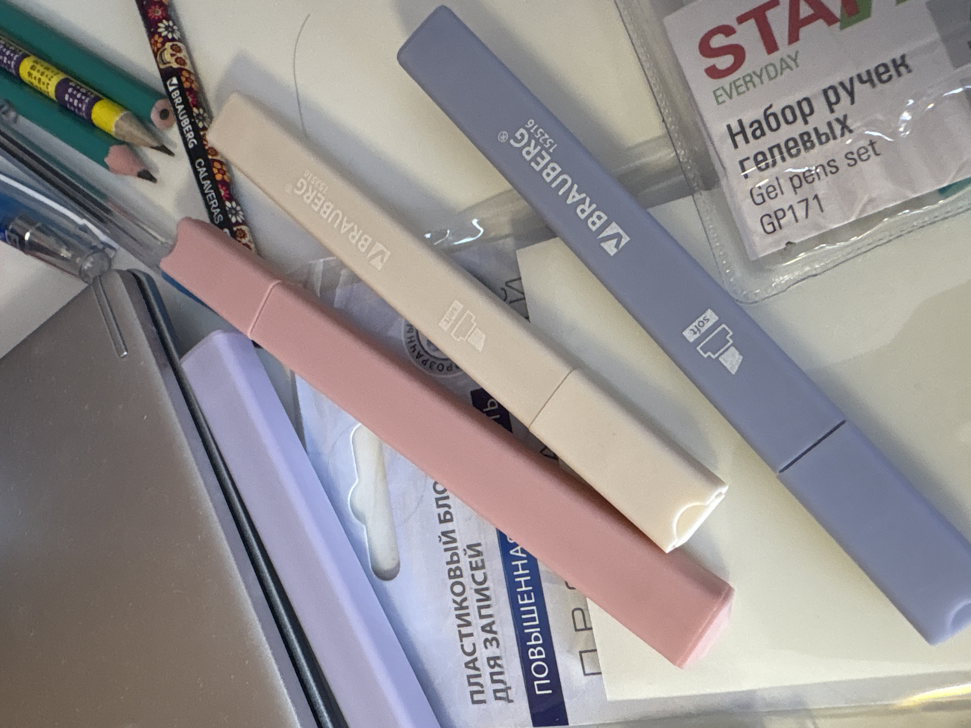 In an indoor setting, possibly a desk or workspace, an arrangement of stationery and office supplies is captured from a top-down perspective. The scene is dominated by three highlighter pens, arranged diagonally. The uppermost highlighter is a soft pink, the middle one is a pale cream, and the bottom one is a muted lavender. Each highlighter is branded with "BRAUBERG" and a product number, "152516".
Beneath the highlighters, partially obscured, is packaging for a "пластиковый блок для записей" (plastic block for notes) with "ПОВЫШЕННАЯ" (enhanced) printed below. Further down, a clear plastic package contains a "Набор гелевых ручек" (set of gel pens) labeled "EVERYDAY STA" with the product code "GP171".
To the left and upper portion of the image, a silver-toned electronic device, possibly a laptop or tablet, is visible. Next to it, several colored pencils are clustered together, including green and yellow pencils with decorative patterns, and a pencil with a colorful floral design. A clear pen is also present among the pencils. The overall impression is one of organized clutter on a flat surface. The location is described as Georgiyevsk, Russia.