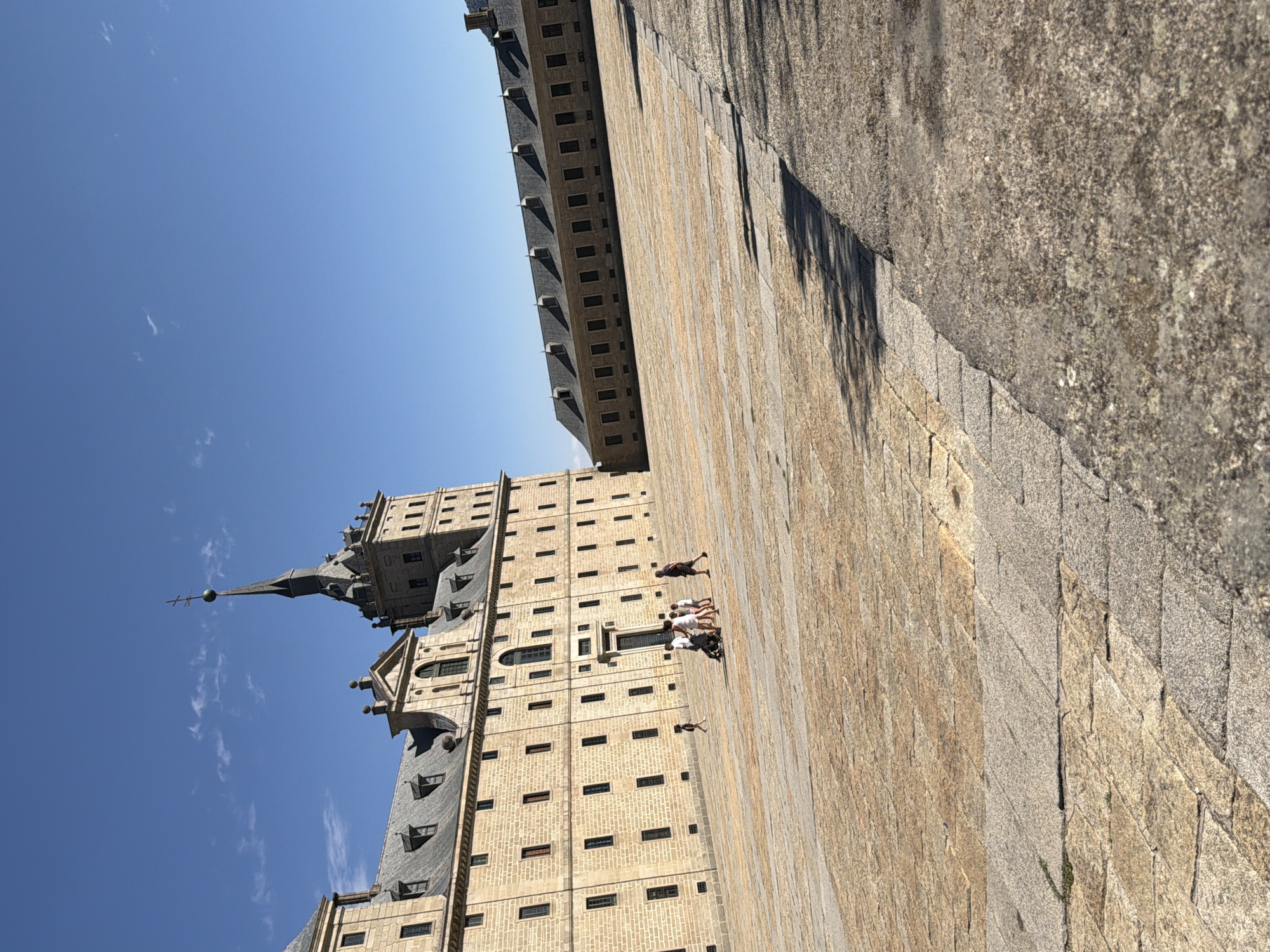 The image shows a wide, sun-drenched plaza in front of the Royal Site of San Lorenzo de El Escorial in San Lorenzo de El Escorial, Spain. The plaza is paved with large, light-colored stone tiles. In the background, a massive, multi-story building with a grey tiled roof and numerous windows dominates the left side of the frame. This building features a tall, dark spire topped with a cross. To the right, another section of the building stretches horizontally, also with a grey tiled roof and evenly spaced windows. Several small figures, appearing to be a family with children and a stroller, are walking across the plaza, casting faint shadows. The sky is a clear, bright blue with a few wispy clouds. The overall scene depicts a bright, sunny day in a historical and architectural complex.