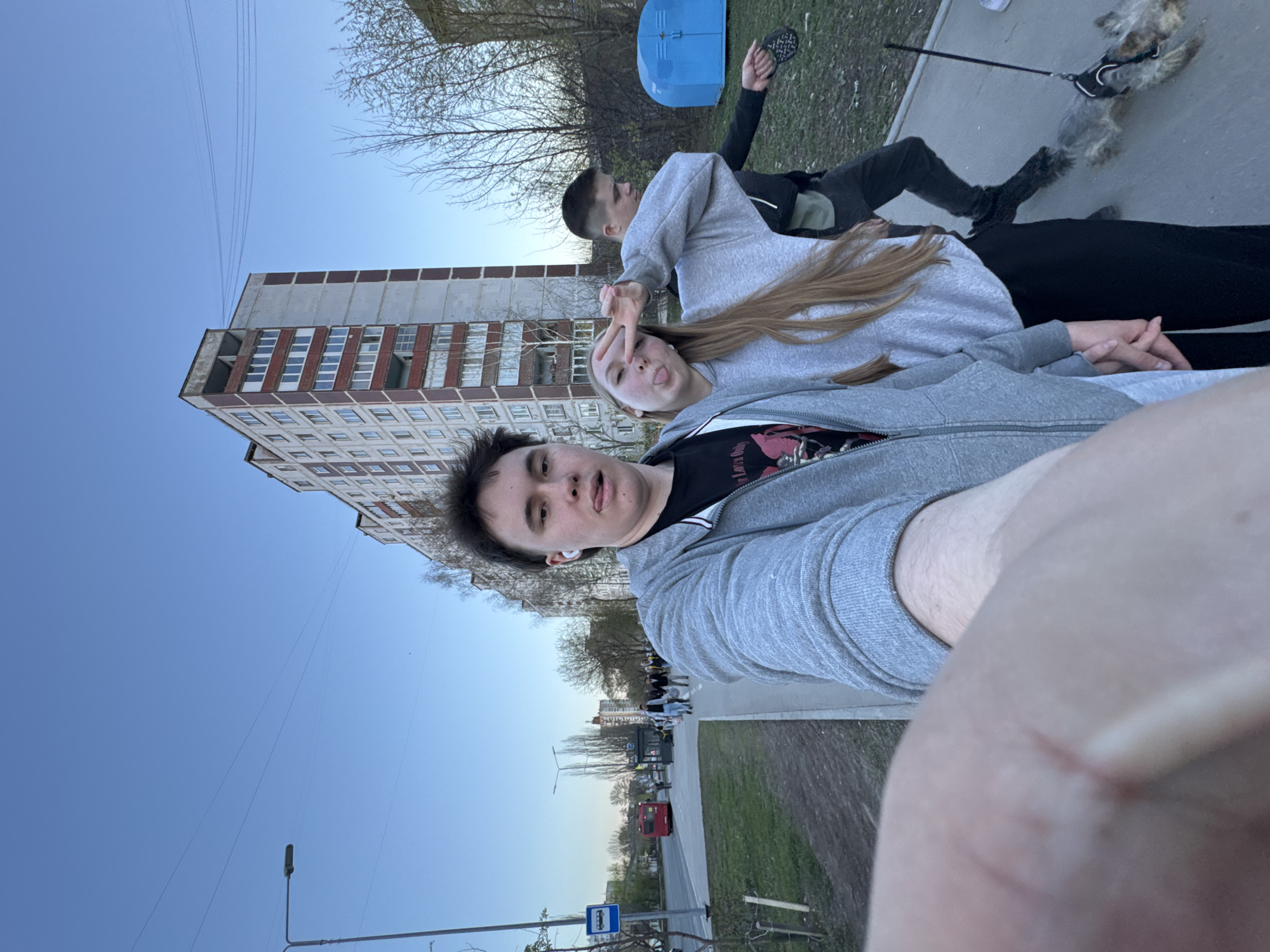 The image shows a selfie of three people, likely young adults, on a sidewalk in Perm, Russia. A partially visible arm and shoulder are in the extreme foreground. A male person with dark hair is looking at the camera. Behind him is a female who is holding up her hand with a "peace sign". Behind the female person, another male is laying face down on the grass. A dog is tied to the male. A tall, Soviet-era apartment building is in the background. A street is to the left. Visible elements include a bus stop sign, a red vehicle on the street, and a blue portable toilet on the grass. The sky is partially visible and blue.