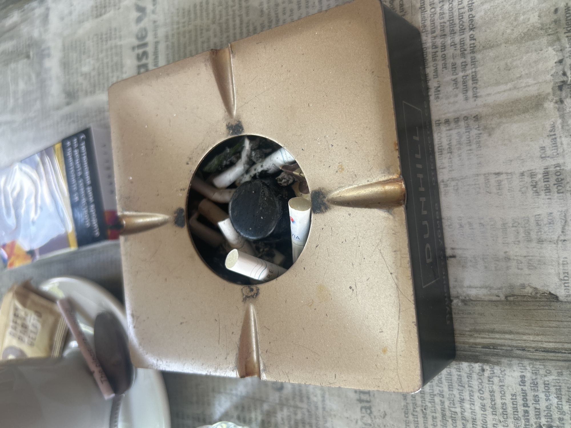 The image depicts a close-up, eye-level view of a brass ashtray filled with cigarette butts, placed on top of a newspaper. The ashtray is square with rounded corners and features four indentations along its rim, likely for resting cigarettes. A black, cylindrical object, possibly a small lid or cap, rests in the center of the ashtray. The newspaper's text is partially visible, with some words and phrases in French and what appears to be Cyrillic script, suggesting an international context. To the left of the ashtray, a white teacup and saucer are visible, with a spoon resting on the saucer. A pink cigarette with visible text is placed on the spoon. The overall scene suggests a casual setting, possibly a cafe or a home, where smoking has occurred. The presence of both French and Cyrillic text could indicate a location in or influenced by both cultures, though without further context, pinpointing a specific location like Plovdiv, Bulgaria is speculative. However, given the presence of Cyrillic script, Plovdiv, being in Bulgaria, is a plausible, though not definitive, location. The lighting is somewhat dim, with a soft, diffused light source. The focus is sharp on the ashtray and its contents, with the background slightly blurred.