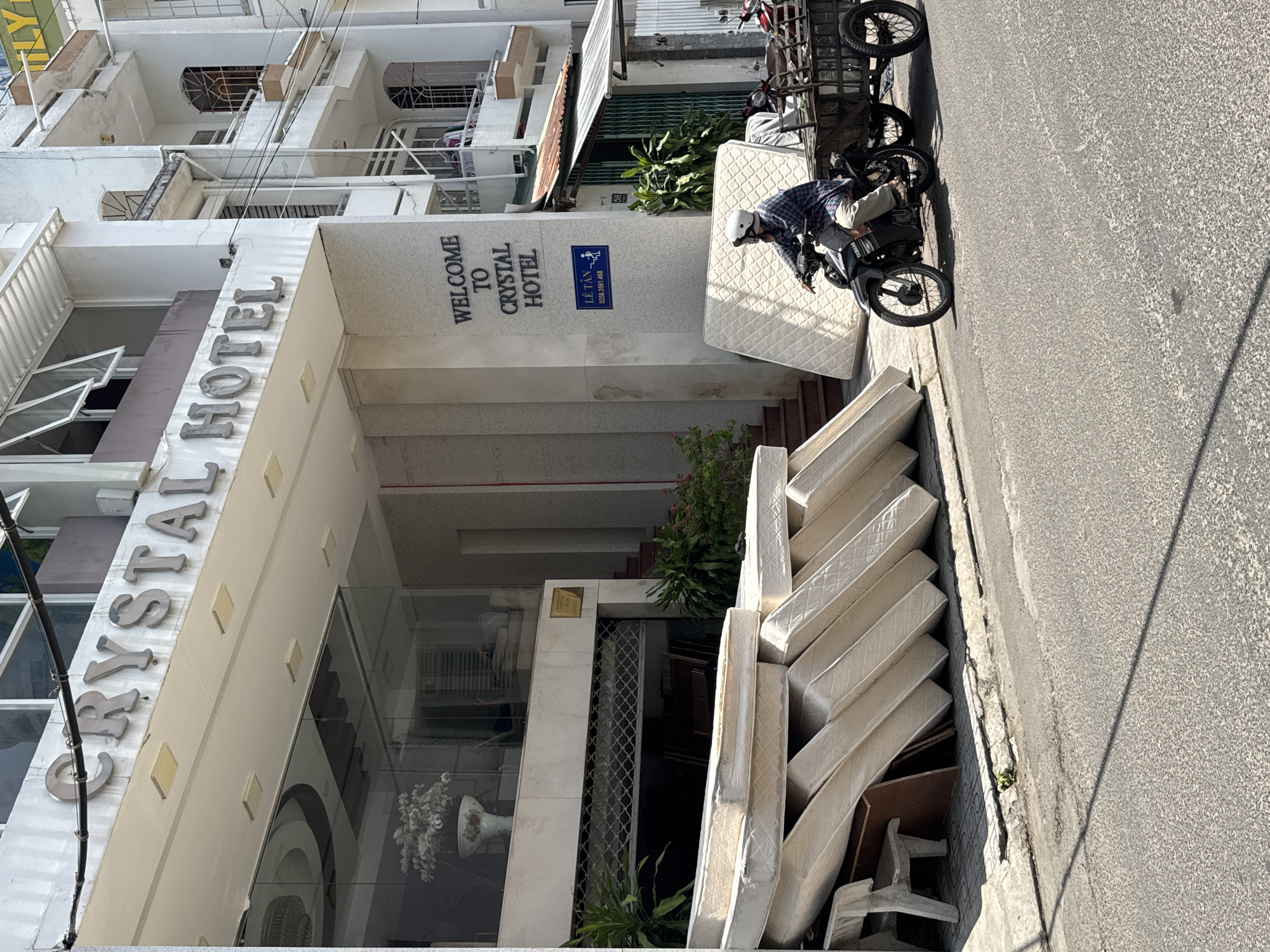 The image depicts the exterior of the "CRYSTAL HOTEL" in Nha Trang, Vietnam. Several mattresses are stacked precariously on the sidewalk in front of the hotel entrance. A man wearing a helmet and a checkered shirt is riding a motorcycle with a trailer attached, which appears to be carrying more mattresses. The hotel has a modern facade with large glass windows and the hotel's name displayed prominently in metallic letters. To the right of the hotel entrance, a blue sign with text and an icon is visible. The street in front of the hotel is paved, and shadows from nearby structures fall across it. The overall scene suggests a commercial area with a focus on hospitality and potentially a mattress removal or delivery operation.