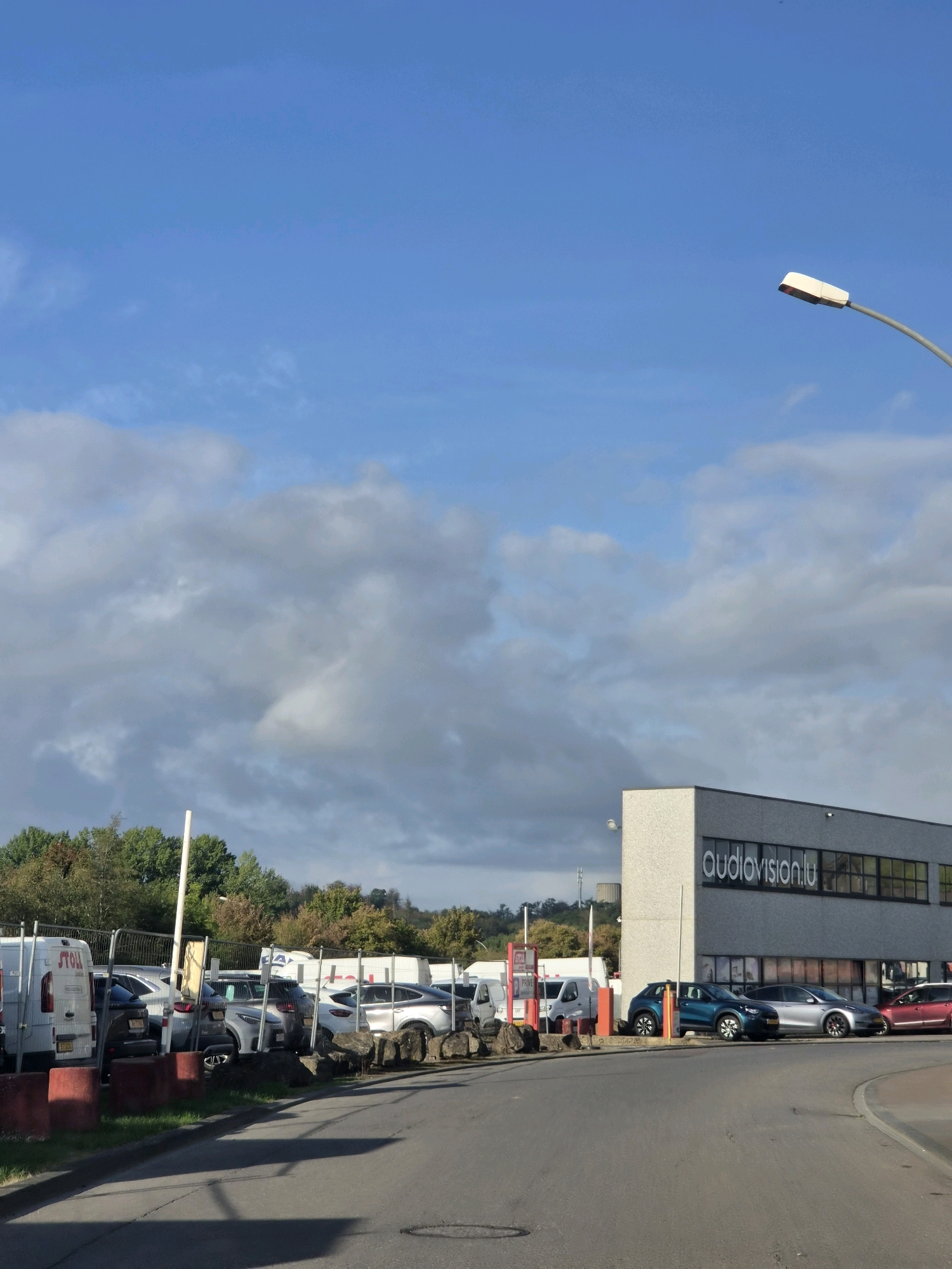 The image depicts a street scene in Leudelange, Luxembourg, under a partly cloudy sky. In the foreground, a paved road curves to the right. Adjacent to the road is a parking lot filled with various vehicles including vans and cars. Behind the parking lot is a modern, grey building with large windows. The building has the text "audiovision.lu" prominently displayed on its facade. A streetlamp is visible in the upper right portion of the image. The environment suggests a commercial or industrial area.
