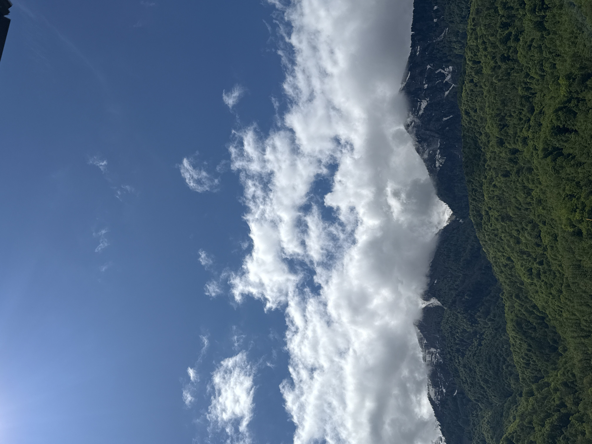 The image presents a view of mountainous terrain under a bright blue sky with scattered clouds. Lush, green vegetation covers the slopes on the right side of the frame, while a darker, rocky mountain face, partially obscured by clouds, is seen to the left. The presence of clouds suggests the altitude. Without additional information or closer details in the image, it is impossible to determine the exact location within Krasnaya Polyana, Russia, specifically.