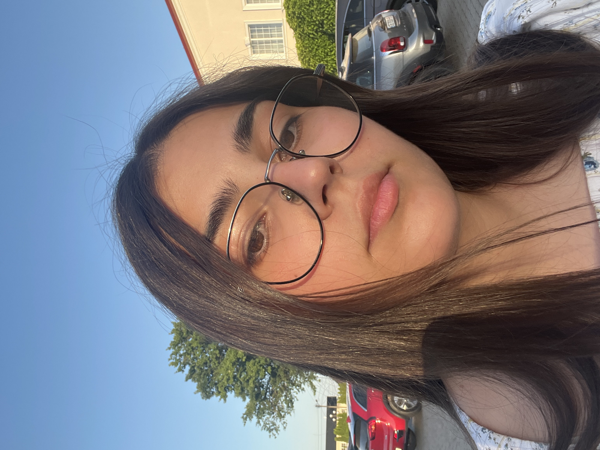 This is a portrait-style selfie taken outdoors in Dubivtsi, Ukraine. The primary subject is a young woman with long, dark hair and glasses. She is looking directly at the camera, and her face is angled slightly, with her lips slightly parted. Her expression appears neutral or perhaps a little playful. She is wearing a light-colored top with a floral pattern.
In the background, there are several cues that indicate the setting and time of day. A building with windows and an ivy-covered wall is visible, along with parked cars. One car is a silver SUV, and another is a red hatchback. The clear blue sky suggests it is daytime, and the warm lighting on the woman's face indicates it might be late afternoon or early evening. The presence of trees further reinforces the outdoor setting. There is no visible text in the image.