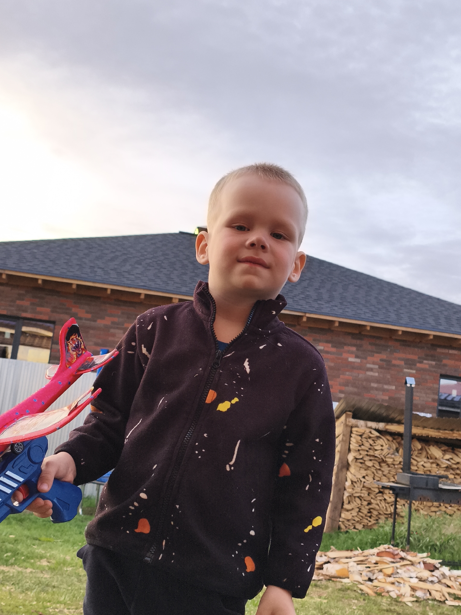 A young, light-skinned child stands outdoors in what appears to be a residential area. He has short blond hair and is wearing a dark zip-up jacket with colorful paint splatters. In his left hand, he holds a toy airplane launcher, featuring a blue handle and a pink plane with designs.
Behind the child is a brick building with a dark-colored roof. To the right, stacked firewood and a barbecue grill are visible. The sky is overcast.
The location is Русский Вожой, Russia, as per the prompt.