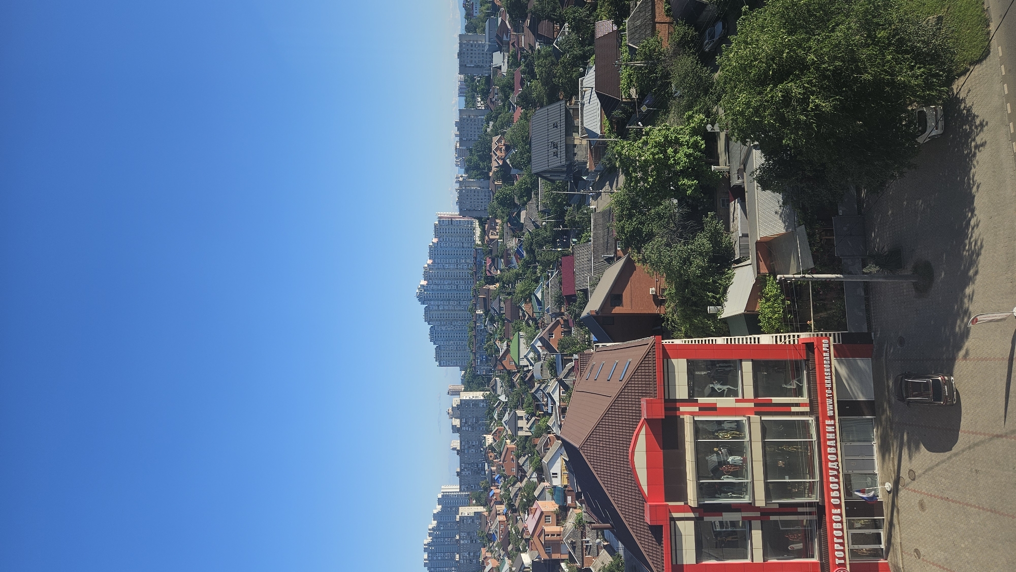 This is a high-angle, outdoor, vertical, full shot of a city landscape under a clear blue sky. In the foreground, a red and white building with large glass windows displays mannequins. The building has signage with Russian text, including "ТОРГОВОЕ ОБОРУДОВАНИЕ" and a website address ending in ".PRO." A dark-colored car is parked in front of the building, and a white car is parked on the right side of the image, partially obscured by a large tree. The middle ground features a dense residential area with numerous houses, many with multi-colored roofs, and abundant green trees. In the background, several tall apartment buildings rise above the lower-lying structures. The scene appears to be in the city of Krasnodar, Russia, given the Cyrillic signage on the foreground building.