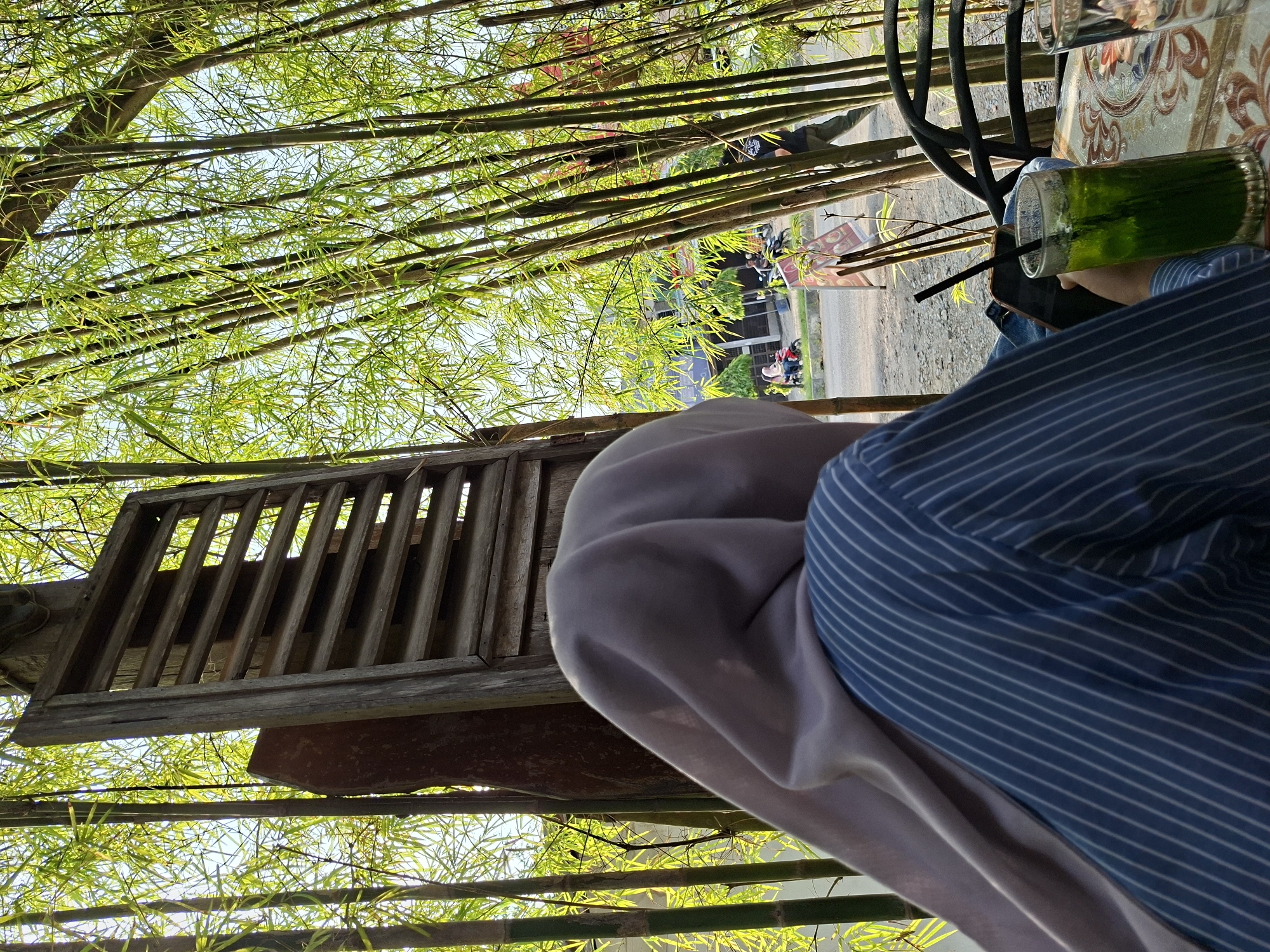 The image shows a scene at what appears to be an outdoor café or restaurant, possibly in Pantegajah, Indonesia. A person in a blue striped shirt and light-colored headscarf is seated at a table, holding a phone and drinking a green beverage with a straw. There are bamboo stalks providing shade or decoration overhead. A wooden structure, like a window or decorative piece, is also visible. In the background, there is a street scene with buildings, motorbikes, and pedestrians. A table with a decorative tablecloth is partially visible, along with a glass.