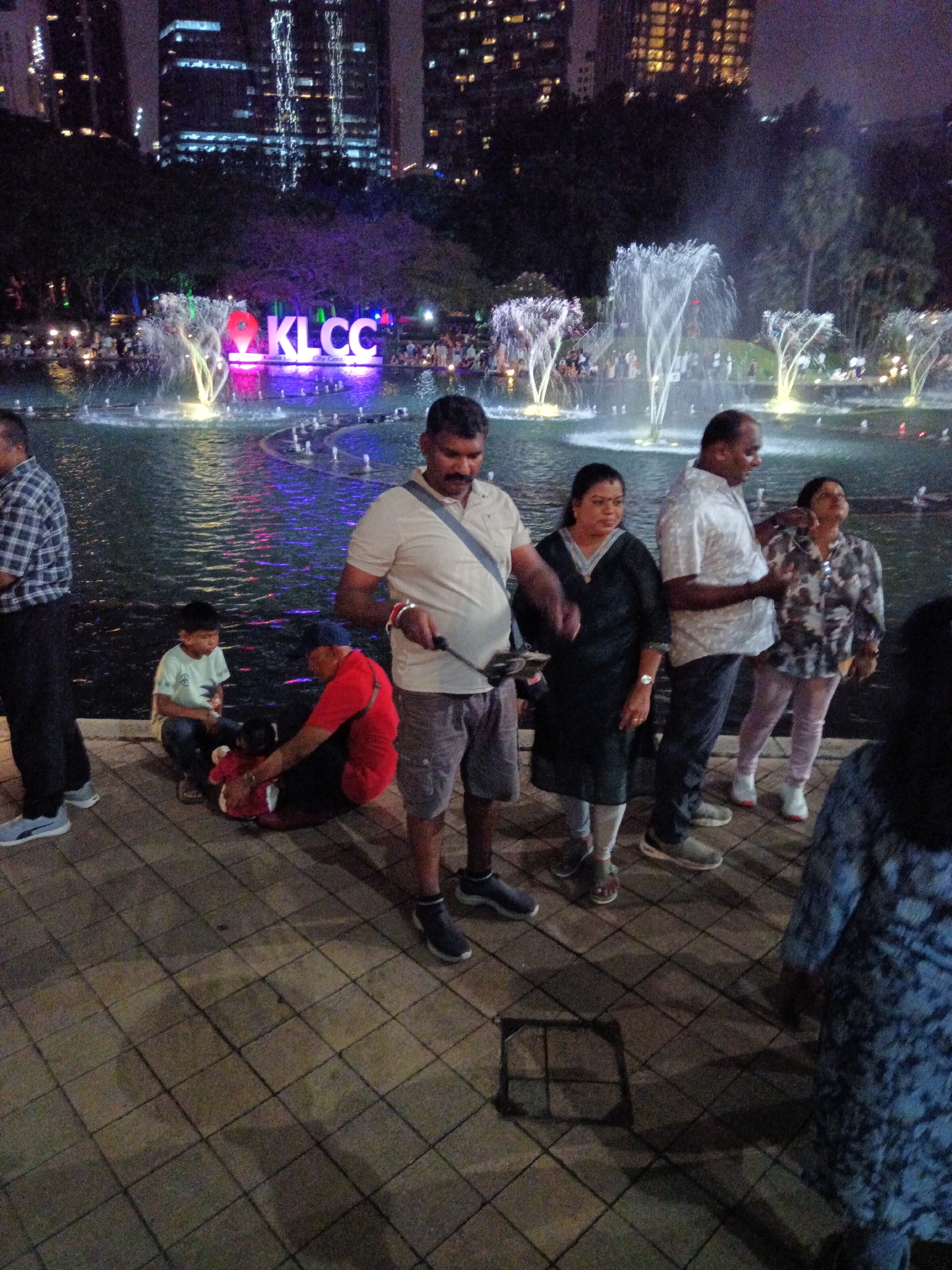 In this nighttime photograph, a group of people are gathered near a fountain display at the Kuala Lumpur City Centre (KLCC) park. The iconic "KLCC" sign, illuminated in pink and purple, is visible in the background, with water jets from the fountain spraying upwards. Several individuals are standing and interacting, while a child sits on the ground. In the foreground, the tiled pavement of the park is illuminated by ambient light and the reflections from the water. The scene is set against a backdrop of illuminated skyscrapers, characteristic of the Kuala Lumpur skyline.