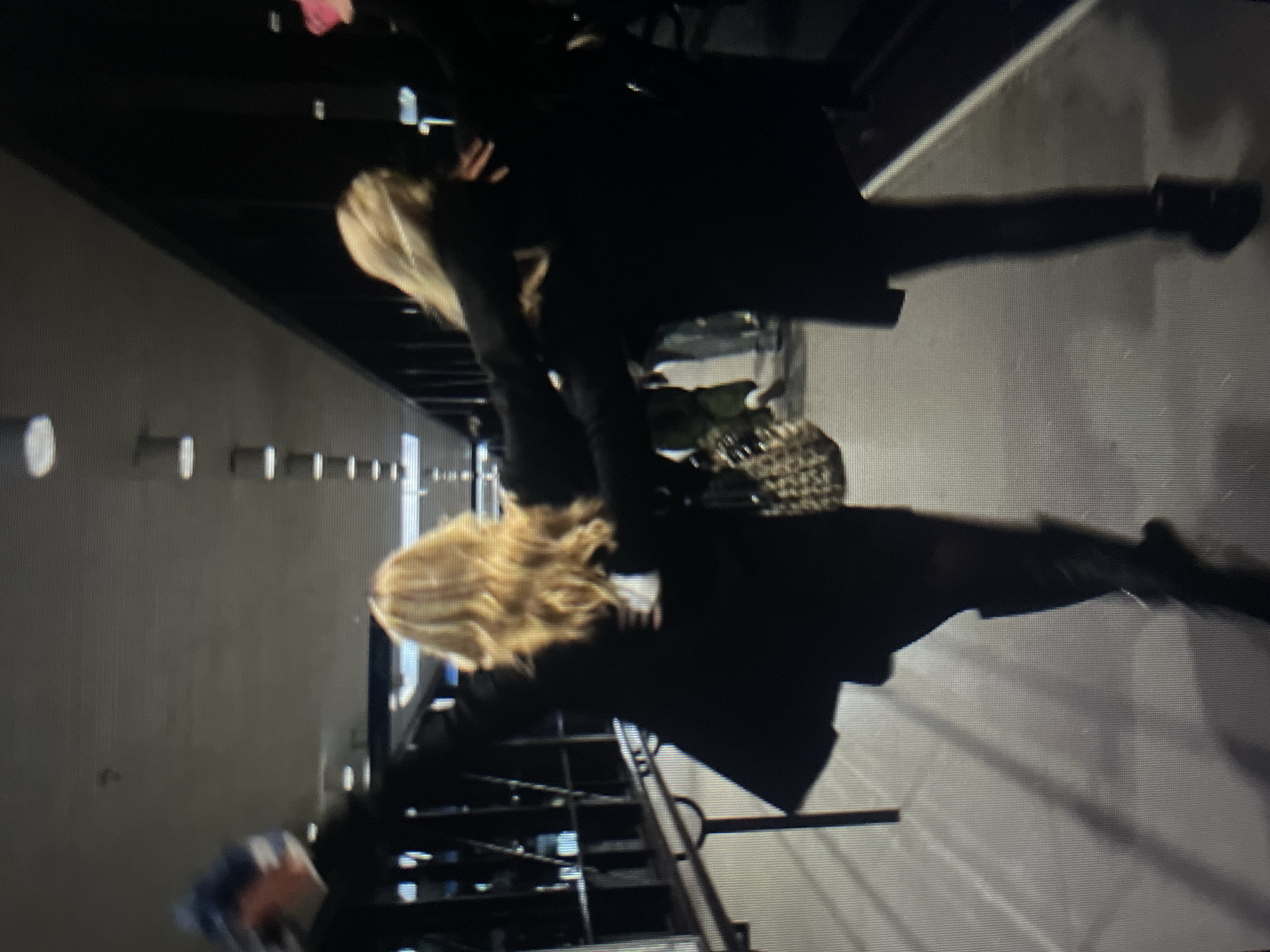 Two individuals are walking away from the camera down a hallway with tiled flooring and overhead fluorescent lighting. The person on the left has long blonde hair, is wearing a black coat, dark tights, and knee-high black boots. They are holding a blue and white object in their raised left hand and have their right arm around the shoulders of the person next to them. The second person on the right is mostly obscured by shadow and the person in front, but appears to have blonde hair and is wearing dark clothing. They are also walking away from the camera, with their left arm around the first person's waist. The environment suggests an indoor public space, possibly a conference center or a large building with an escalator visible in the background. The lighting casts distinct shadows on the floor. The location is identified as Kołobrzeg, Poland.