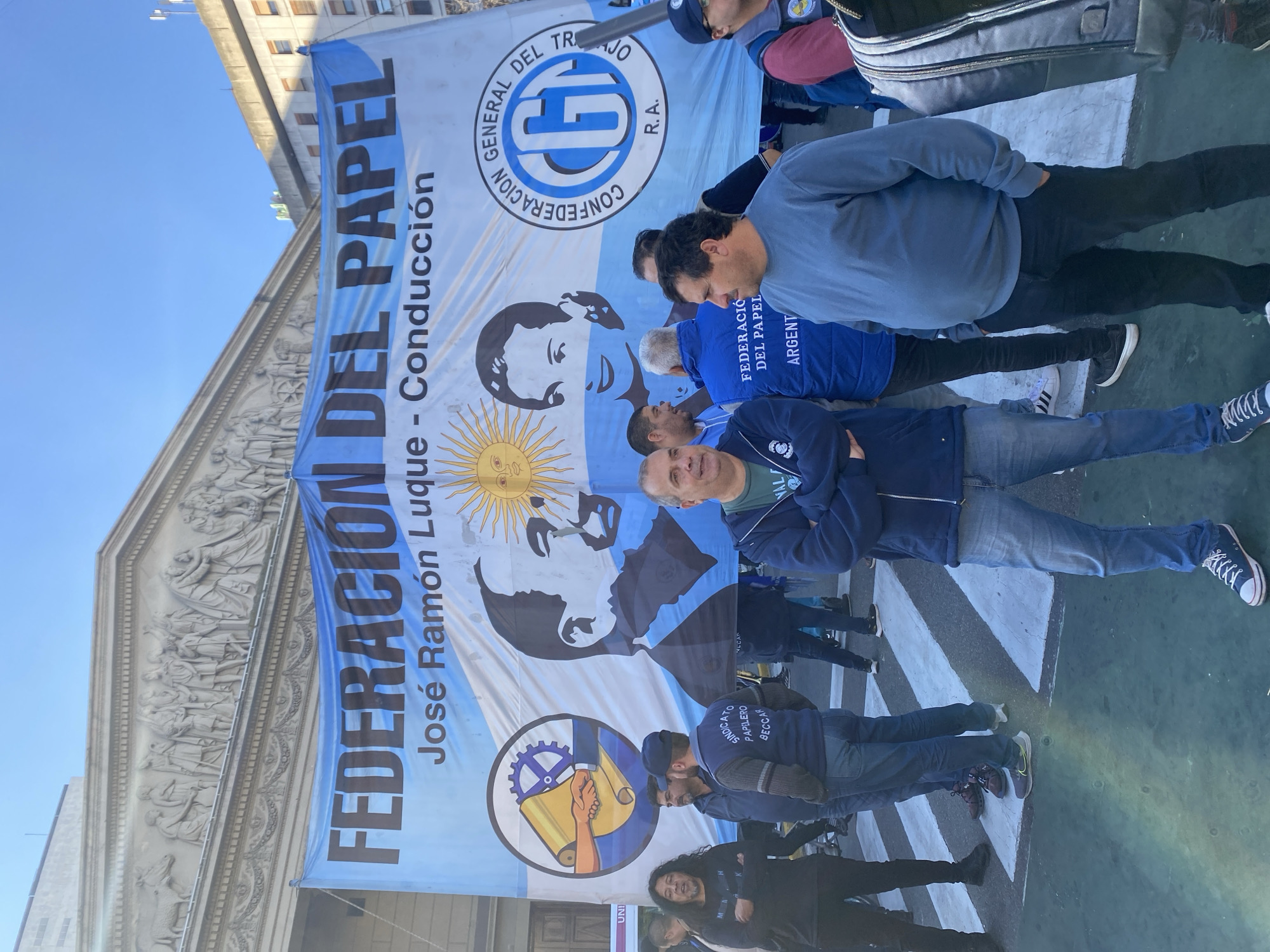 In Buenos Aires, Argentina, a horizontal banner is displayed in front of a building with classical architectural elements. The banner reads "FEDERACIÓN DEL PAPEL" with the text "José Ramón Luque - Conducción" underneath. The flag also features images of two people in black and white, a sun, and the words "CONFEDERACIÓN GENERAL DEL TRABAJO". People stand in front of and behind the banner. The ground is a crosswalk.