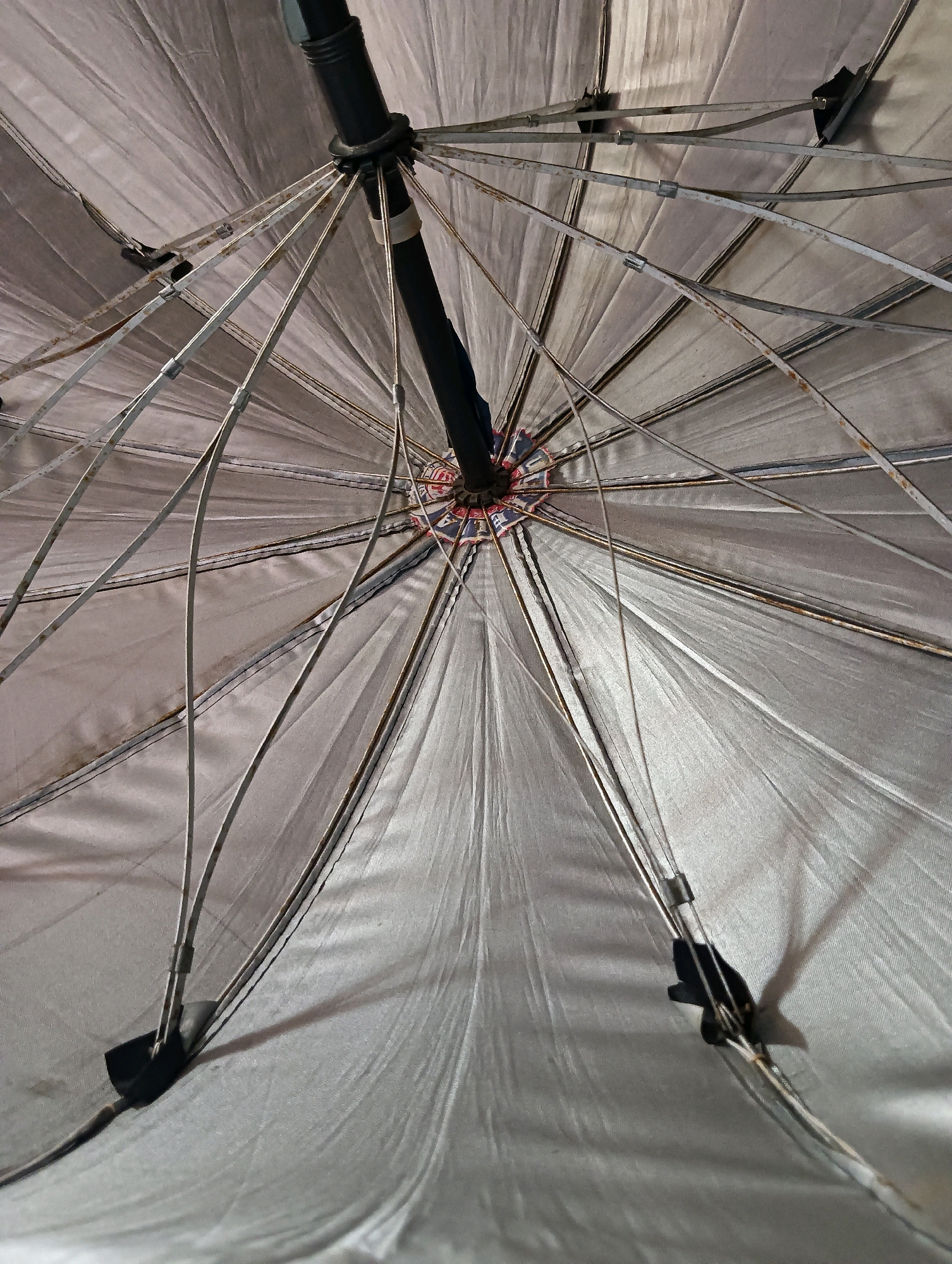 An inside-view photograph of a partially opened, silver-colored umbrella is presented. The umbrella exhibits signs of wear and tear, with visible rust on its metal ribs and some dirt on the fabric. The central pole is black and extends upwards, connecting to a circular mechanism from which numerous metal spokes radiate outwards to support the canopy. These spokes are thin and have small clips attaching them to the fabric. The fabric itself is slightly creased and appears to be made of a thin, somewhat reflective material. The lighting suggests an indoor setting or overcast conditions, casting soft shadows from the umbrella's structure onto its surface. No people are visible in the image. The image does not contain any information that could link it to the city of Hosapete, India.