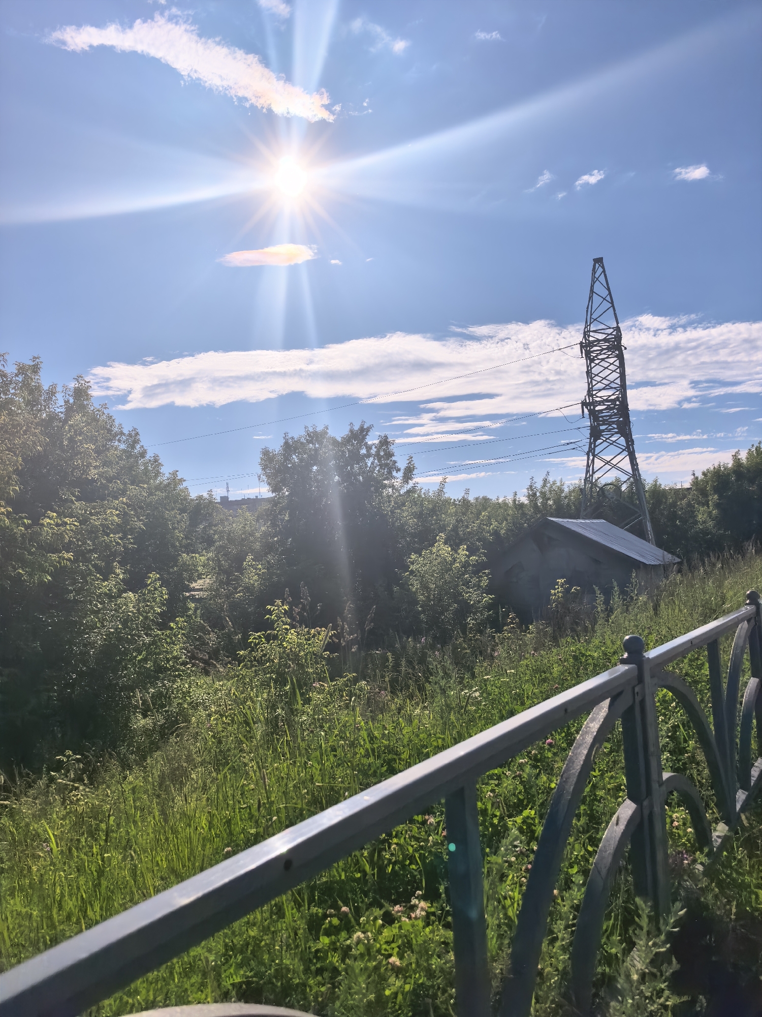 The image shows a bright, sunny day with the sun prominently featured in the upper left quadrant, casting lens flares. The sky is a clear blue with scattered white clouds. In the middle ground, a tall, latticed metal pylon stands amidst lush green trees and vegetation. To the right of the pylon, a small, weathered building with a metal roof is partially visible. In the foreground, a dark metal fence runs diagonally from the bottom left to the right edge of the frame, with green grass and wildflowers visible beyond it. The scene appears to be outdoors, possibly in a park or a rural area with industrial elements.