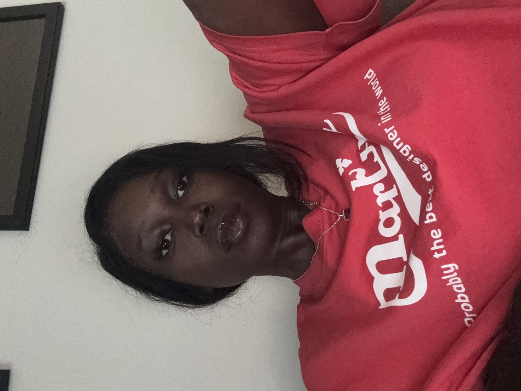 A woman is posing for a selfie indoors, wearing a red t-shirt with white lettering that reads "Markt probably the best designer in the world." She has dark hair and skin, and she is wearing a necklace. There is a black picture frame hanging on the wall behind her. The location appears to be inside a residence. It is not possible to identify the exact location in Los Angeles based on this image.