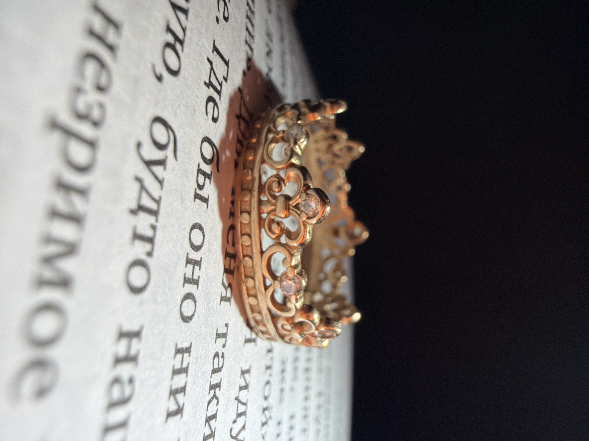 A golden, crown-shaped ring rests on an open page of a book, displaying printed text in black Cyrillic characters. The ring, crafted from gold-toned metal, features intricate filigree scrollwork and several small, clear, faceted stones embedded at its peak points and along its band. The paper is off-white, and the visible text includes partial words such as "Где бы оно ни", "будто", and "незримое". A distinct shadow of the ring is cast onto the page, indicating a directional light source from the upper left. The background is predominantly dark and out of focus, rendering it an indistinct deep black. No individuals are present. The scene is located in the city of Unknown, Unknown.