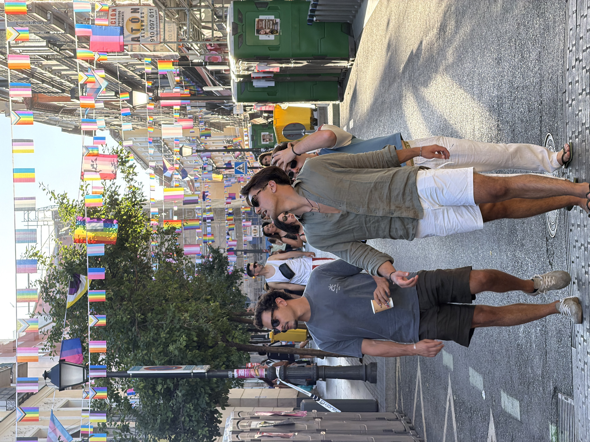 This is a street-level, eye-level, medium shot of three young men walking down a city street. The street is decorated with numerous rainbow-colored flags, indicating a Pride event or celebration. The buildings on either side of the street are multi-story structures, some with balconies and arched windows, suggesting an urban European setting. The sky is clear and bright, indicating daytime.
In the foreground, two men are walking side-by-side. The man on the left is wearing a dark gray t-shirt, dark shorts, and sneakers. He is holding a paper cup in his right hand and has sunglasses on. The man on the right is wearing a khaki-colored unbuttoned shirt over a light-colored t-shirt, white shorts, and sandals. He is looking upwards, with sunglasses on. A third man, wearing a light blue t-shirt and light-colored pants, is walking slightly behind and to the right of the man in the khaki shirt.
In the background, other people are visible walking along the street and on the sidewalks. There are portable toilets on the left side of the street and scaffolding and construction equipment on the right side. A street lamp is visible on the left, and commercial signage is present on the buildings. The street itself is paved with asphalt. The overall atmosphere appears to be festive and lively.
Based on the architectural style of the buildings and the presence of the specific flag decorations, this scene is likely located in the Chueca neighborhood of Madrid, Spain, known for its LGBTQ+ community and vibrant street life, particularly during Pride events.
